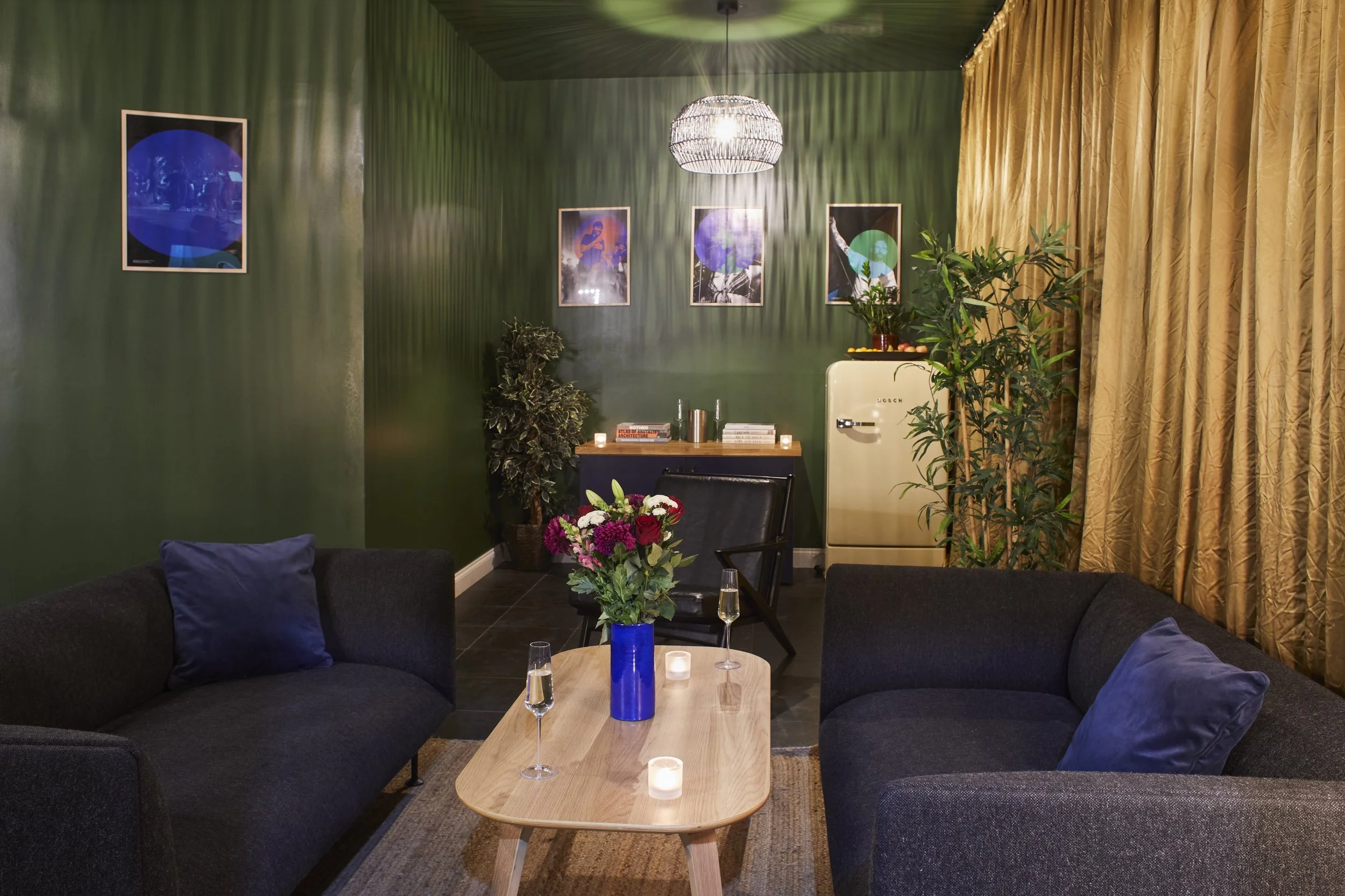 Living room with green walls, black sofas with blue pillows, wooden coffee table with candles and flowers, potted plants, gold curtains, paintings on the wall, and a small vintage fridge.