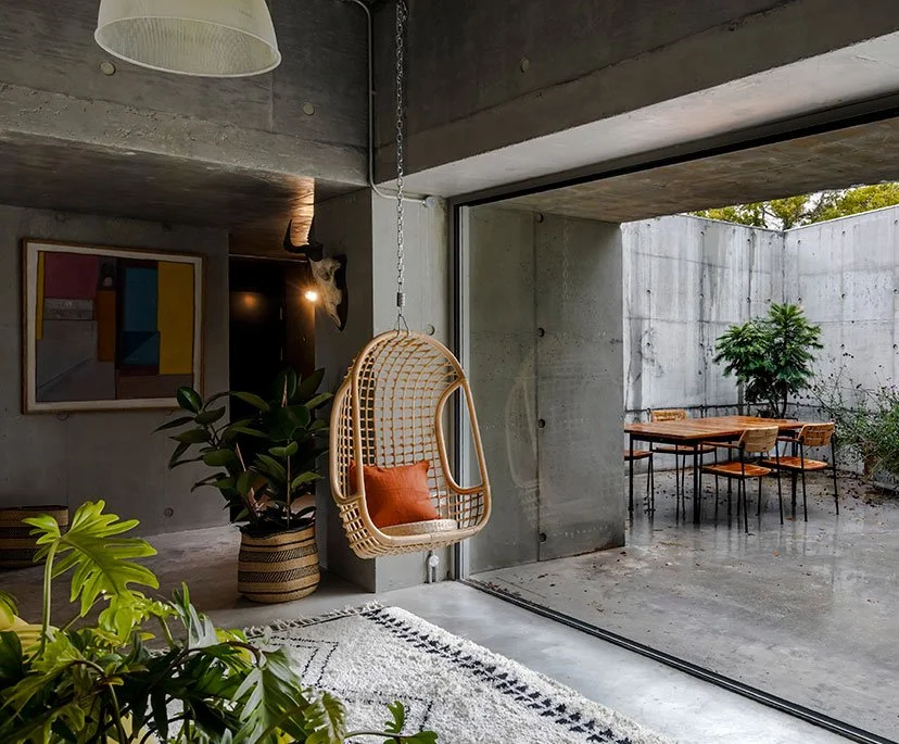 Modern indoor living space with a hanging rattan chair, a large potted plant, and a sliding glass door leading to an outdoor patio with a table and chairs, and a small tree.