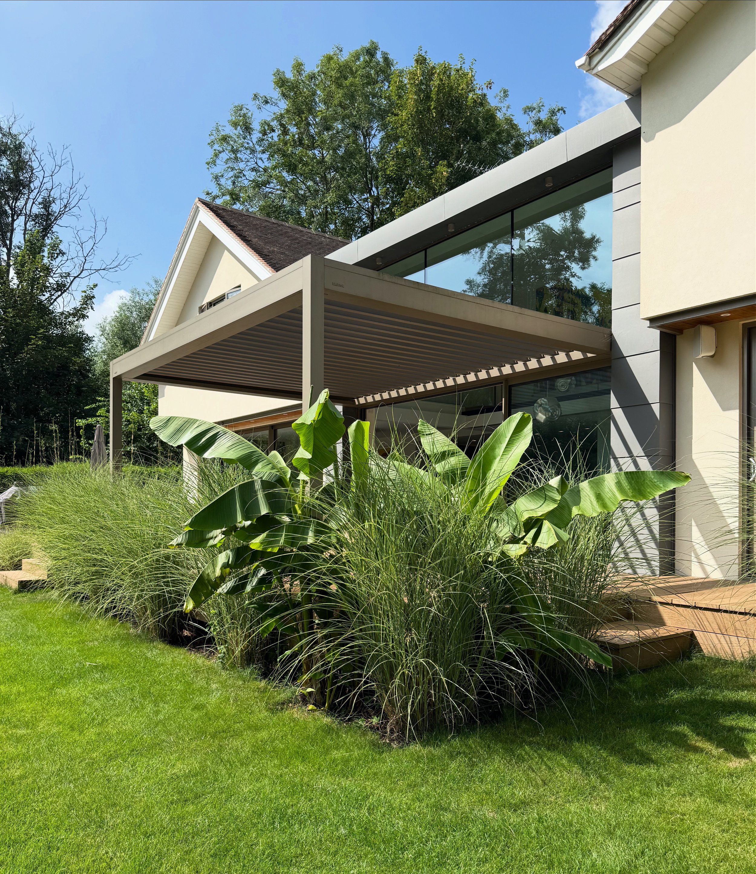 Modern house with large glass windows and a garden with lush green plants, including banana plants, under a partly cloudy sky.