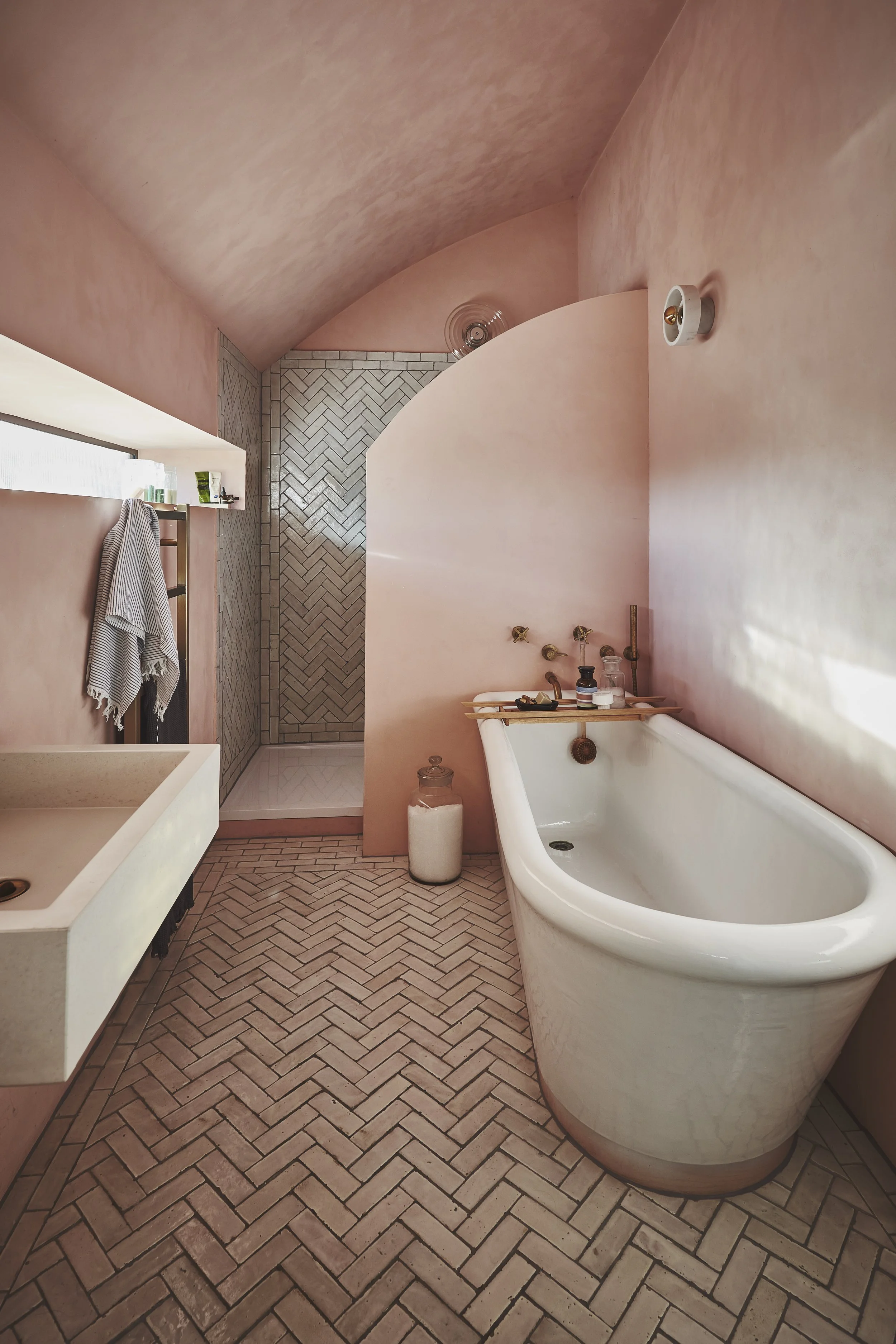 A bathroom with pink walls and herringbone patterned tile flooring. Contains a white bathtub, a small rectangular sink, a window with a towel hanging, a shower area with herringbone tiles, and various small toiletries on a tray and ledge.