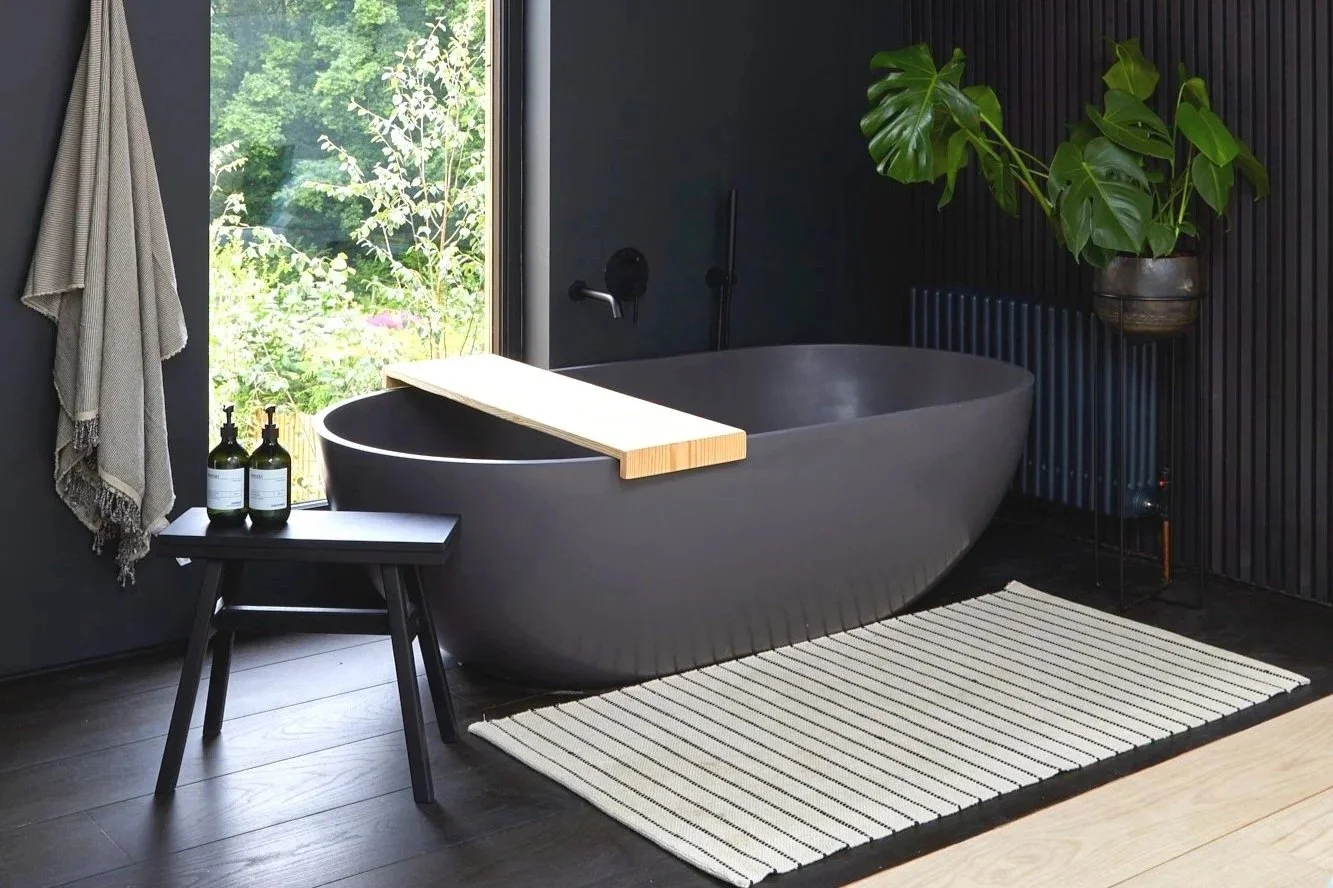 Modern bathroom with a black bathtub, large window, green plant, side table with two bottles, striped rug, and black wooden wall paneling.