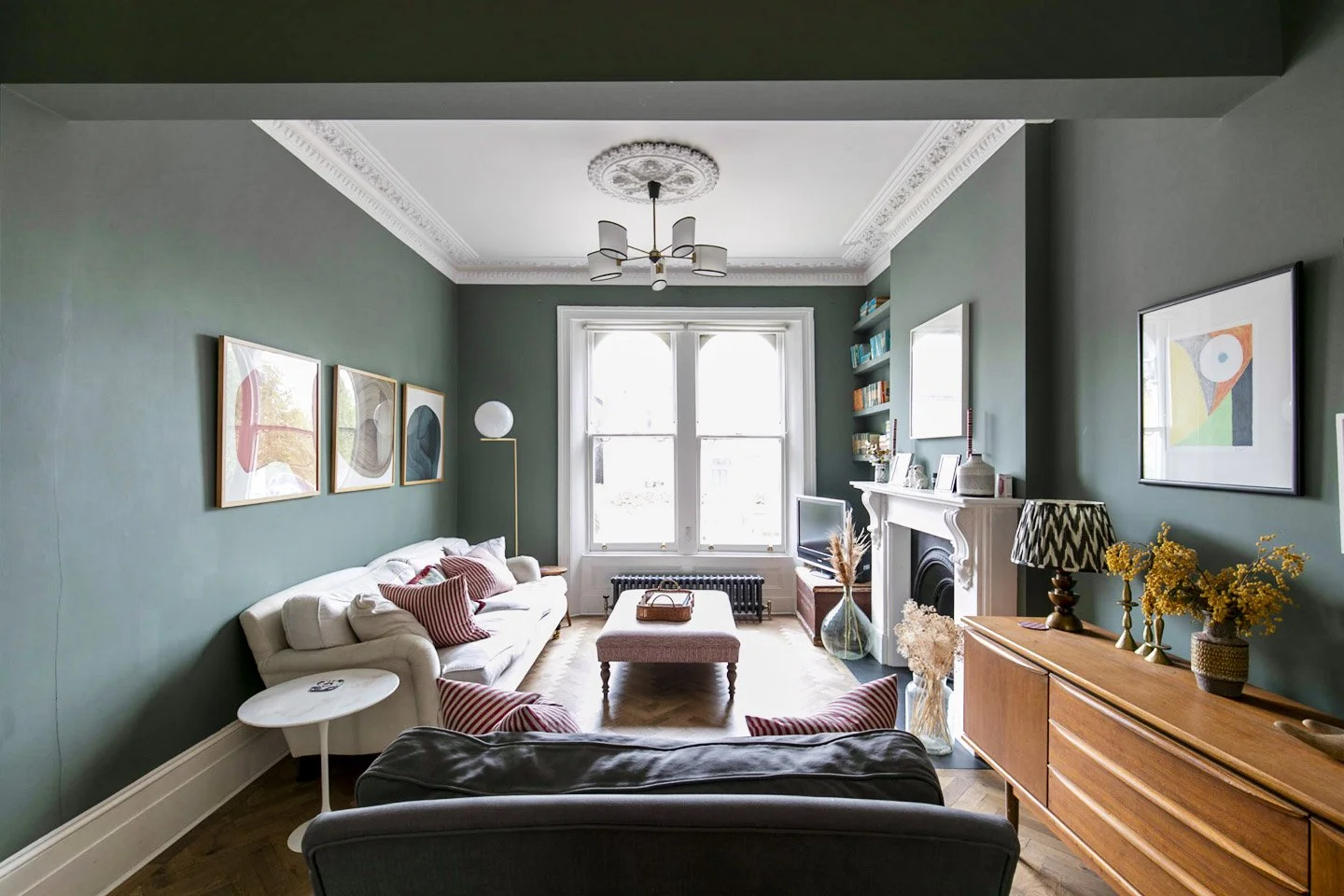 Living room with green walls, white sofa with red and white striped pillows, pink ottoman, wooden sideboard with yellow flowers, fireplace, bookshelves, framed artwork, window, and modern chandelier.