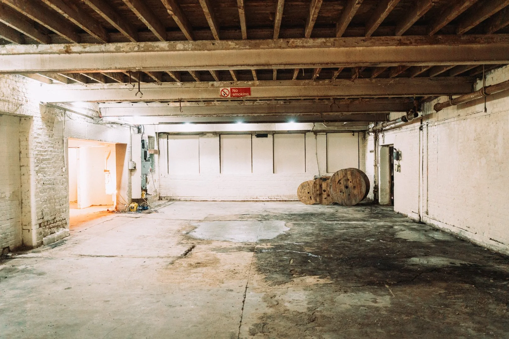 Empty industrial garage with exposed brick walls, concrete floor, large wooden spools, pipes, and electrical boxes, illuminated by overhead lights.