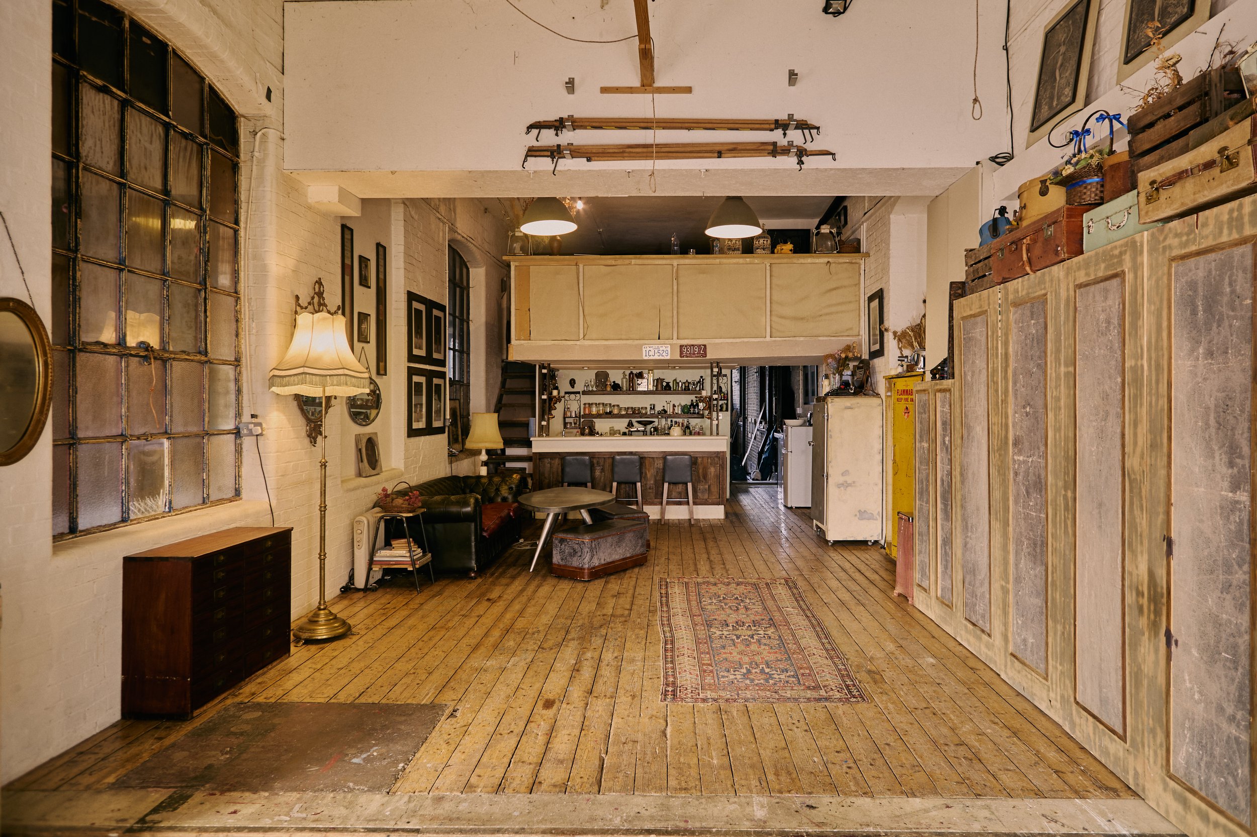 Vintage-style living room with wooden floors, white brick walls, and large industrial windows. Contains a leather sofa, floor lamps, small tables, and a bar area in the background. Decorative items and framed pictures adorn the walls.