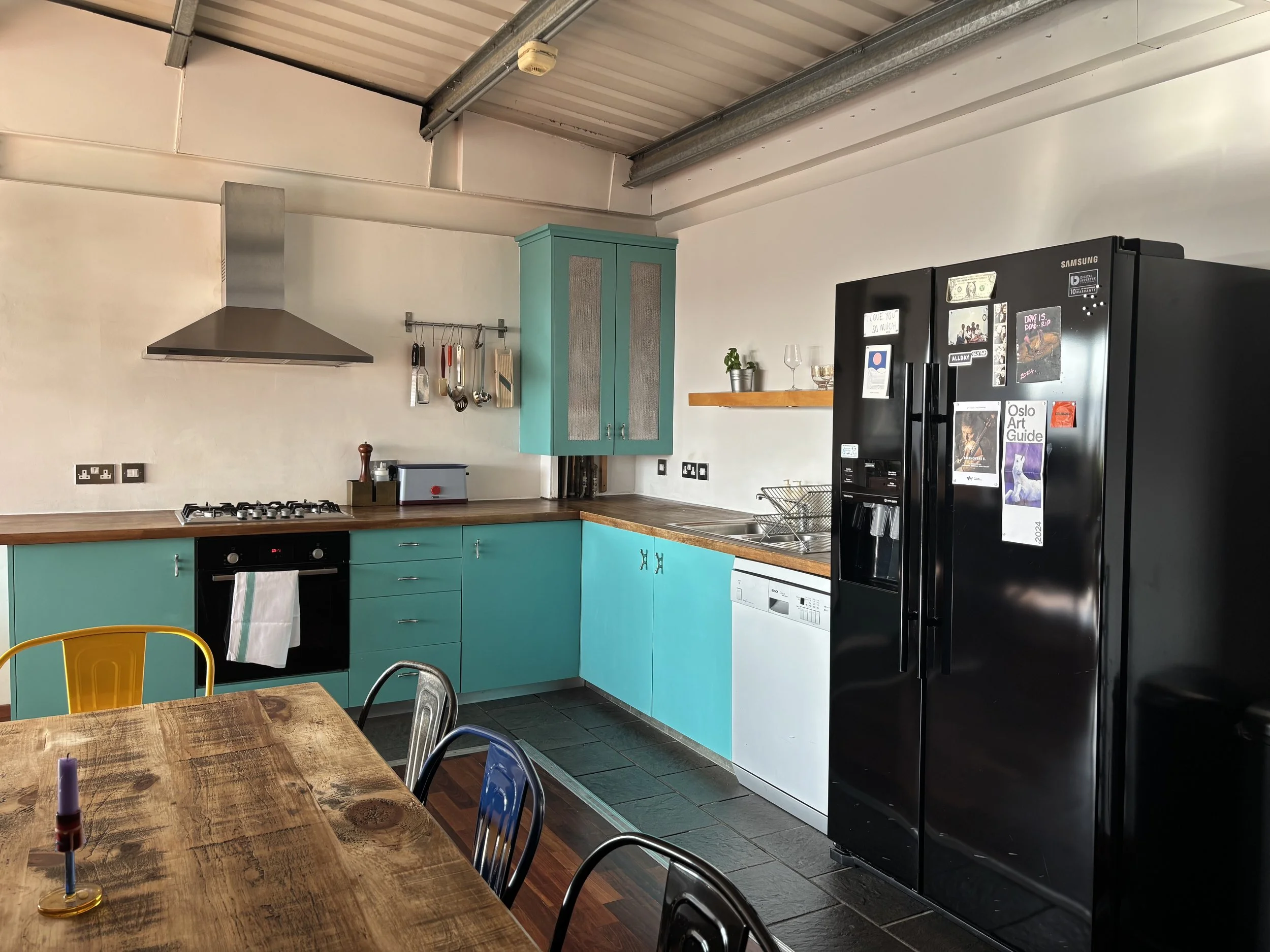 A modern kitchen with turquoise cabinets, a black refrigerator with magnets, a wooden table with chairs, a stove, a sink, and hanging utensils.