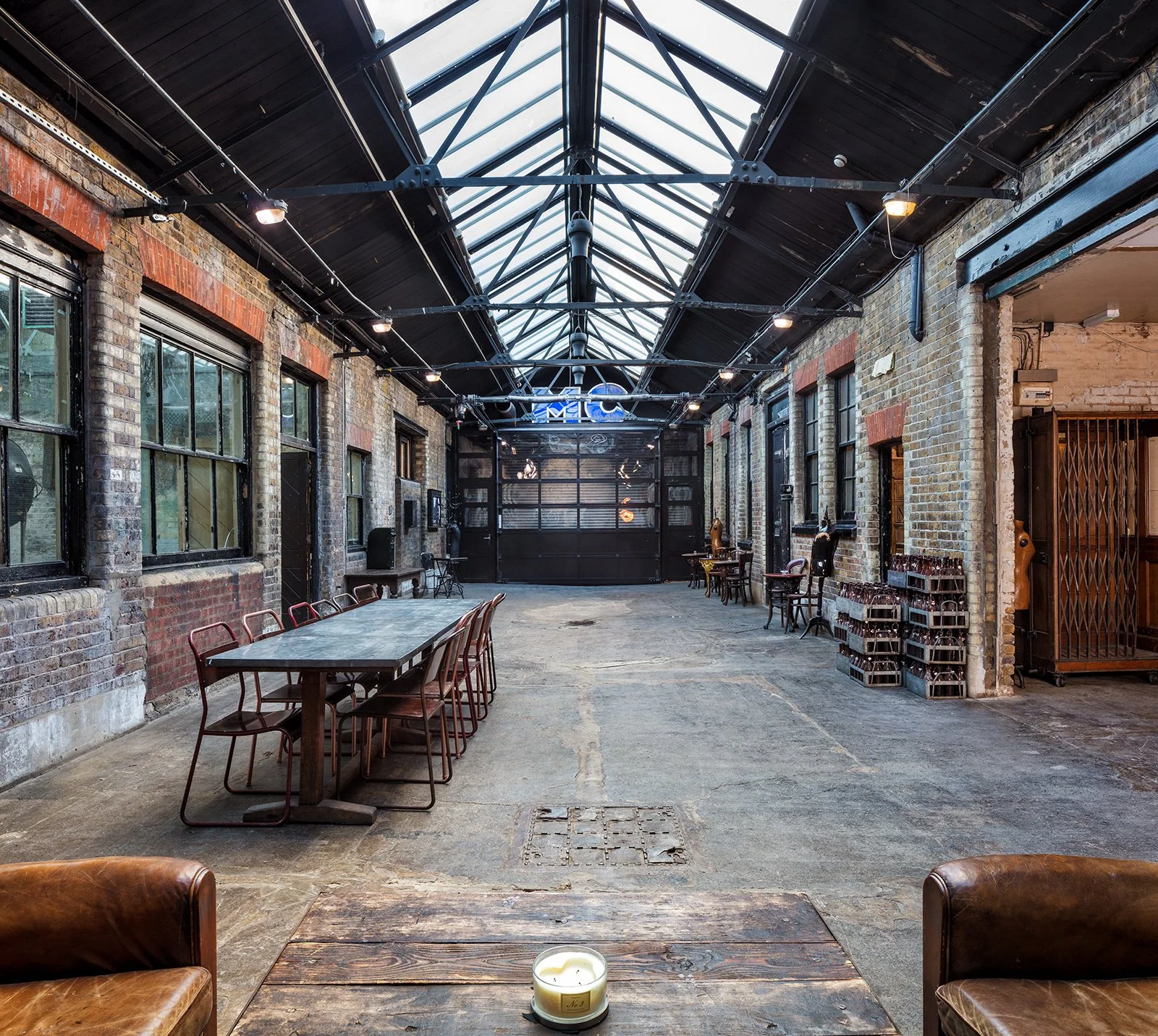 Indoor industrial-style space with exposed brick walls, large windows, and a glass vaulted ceiling, featuring a long table with chairs, small tables and chairs along the walls, and a candle on a wooden table in the foreground.