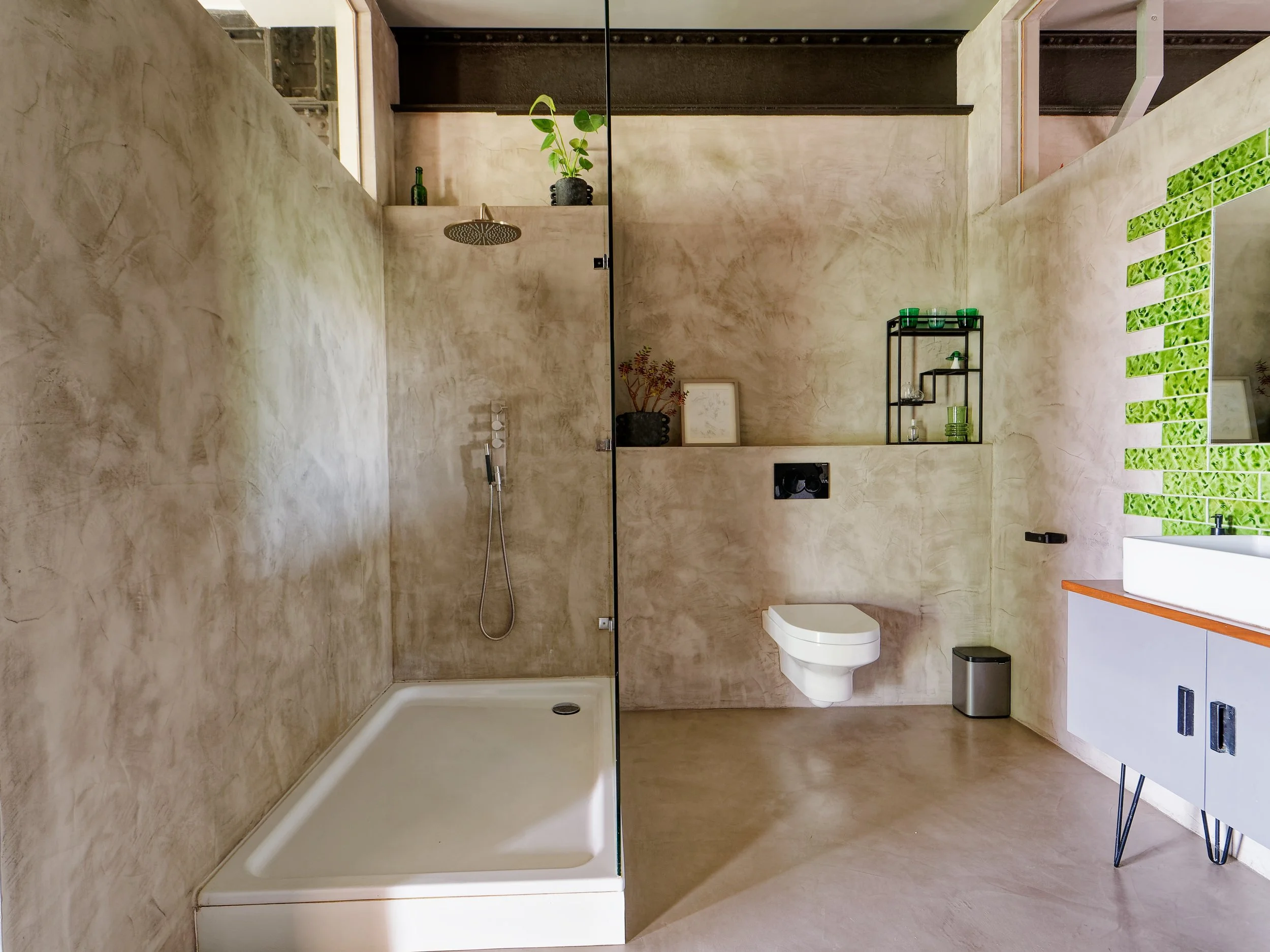 Modern minimalist bathroom with beige textured walls, a walk-in shower with a rain showerhead, a wall-mounted toilet, a black shelving unit with green glasses and decor, a white vanity with a vessel sink, and a green tile accent wall.