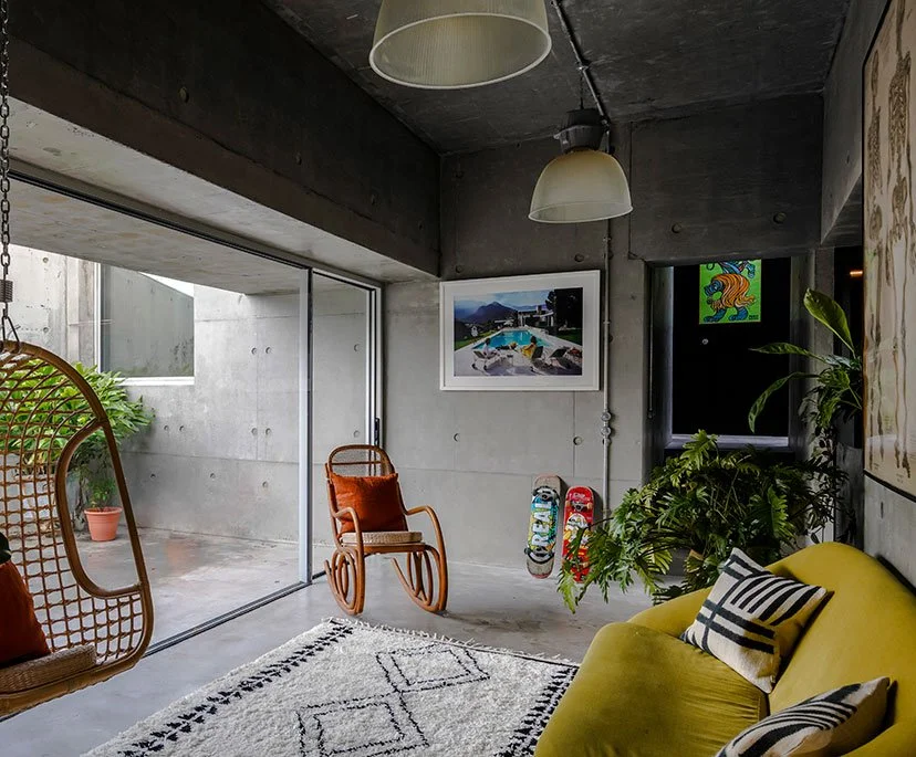 Modern living room with concrete walls, sliding glass door, hanging chairs, skateboard on wall, yellow couch with black and white pillows, indoor plants, colorful artwork.