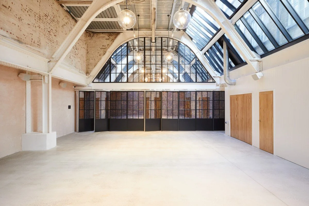 Empty industrial-style room with high vaulted glass ceiling, hanging light fixtures, exposed brick wall, and wooden doors.