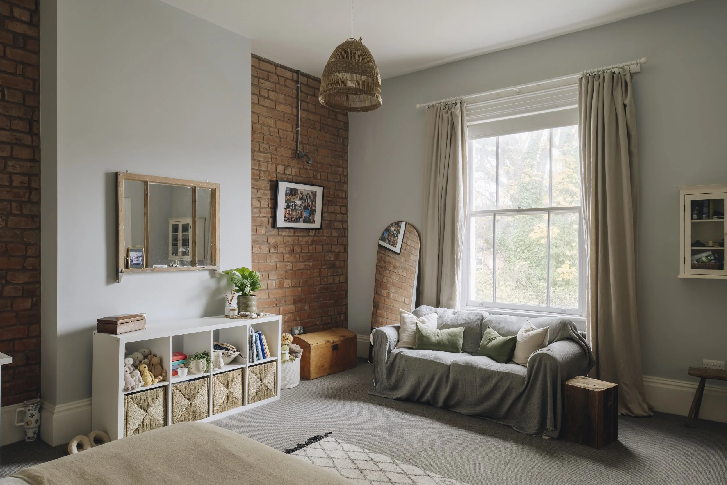 Cozy living room with a large window, brick accent wall, white shelves with toys and books, a mirror, and a sofa with green and beige pillows.