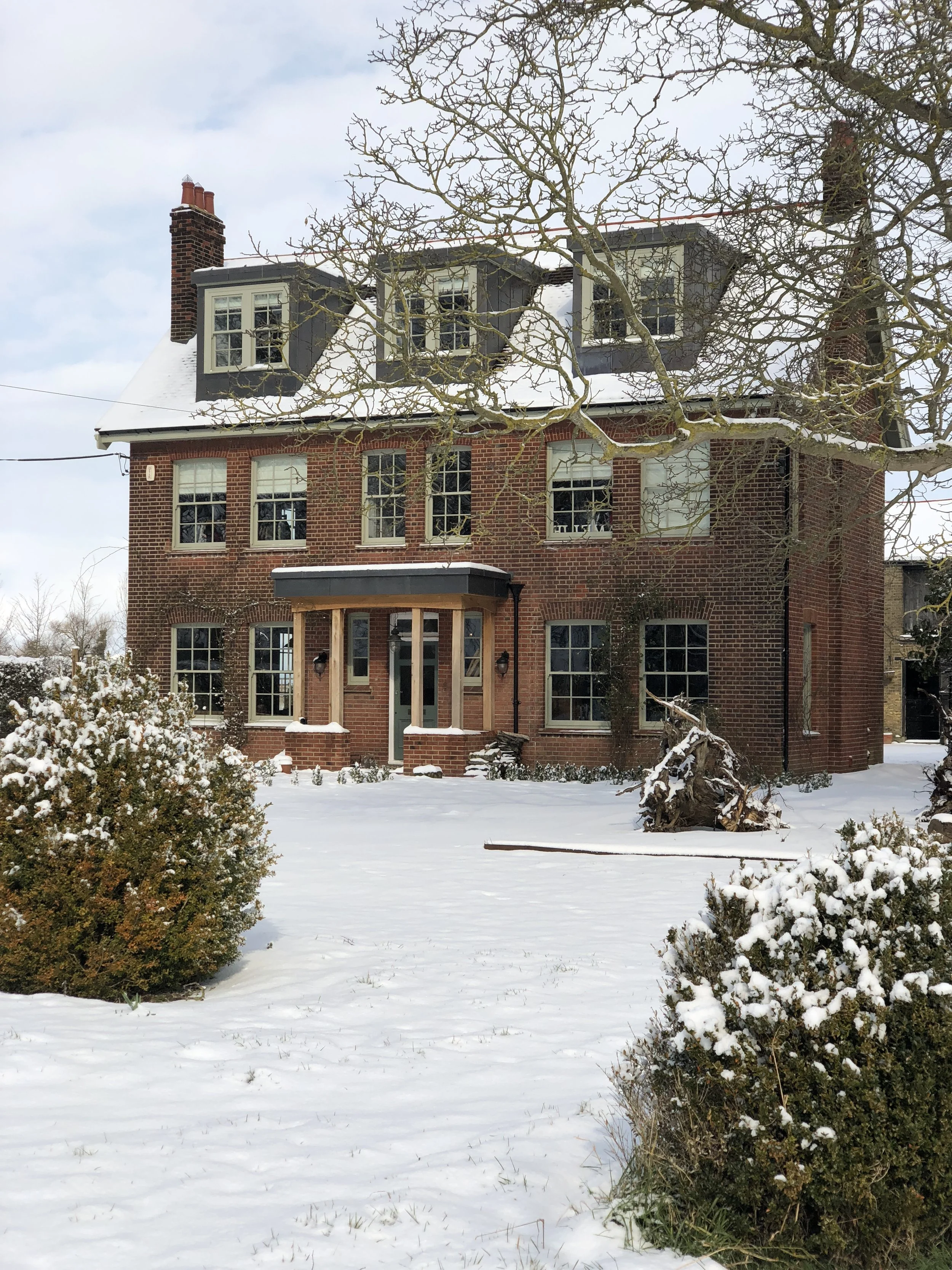 A multi-story brick house with snow on the roof and in the yard, leafless trees in front, and bushes partly covered in snow.