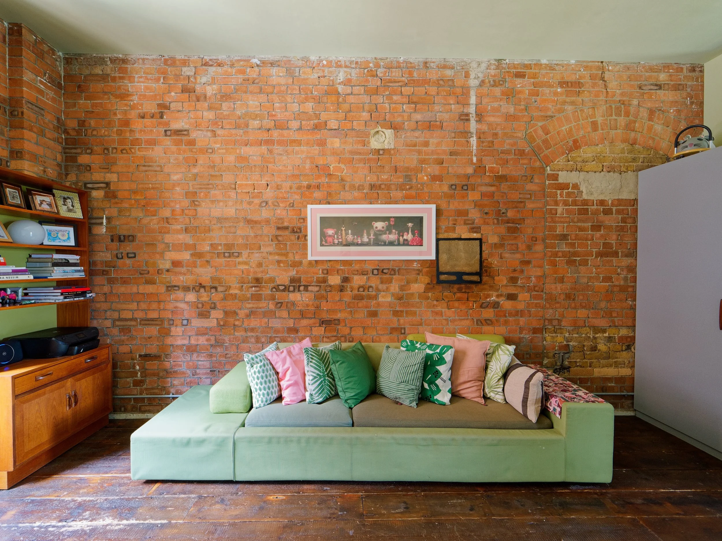 Living room with a green sofa, colorful pillows, a brick wall background, a wooden shelf on the left with decor and books, and a framed picture on the brick wall.