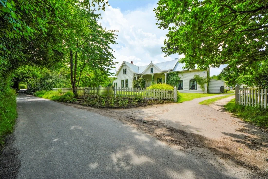A white house with a gray roof surrounded by greenery and a white picket fence, with a dirt driveway and a paved road nearby, under a partly cloudy sky.