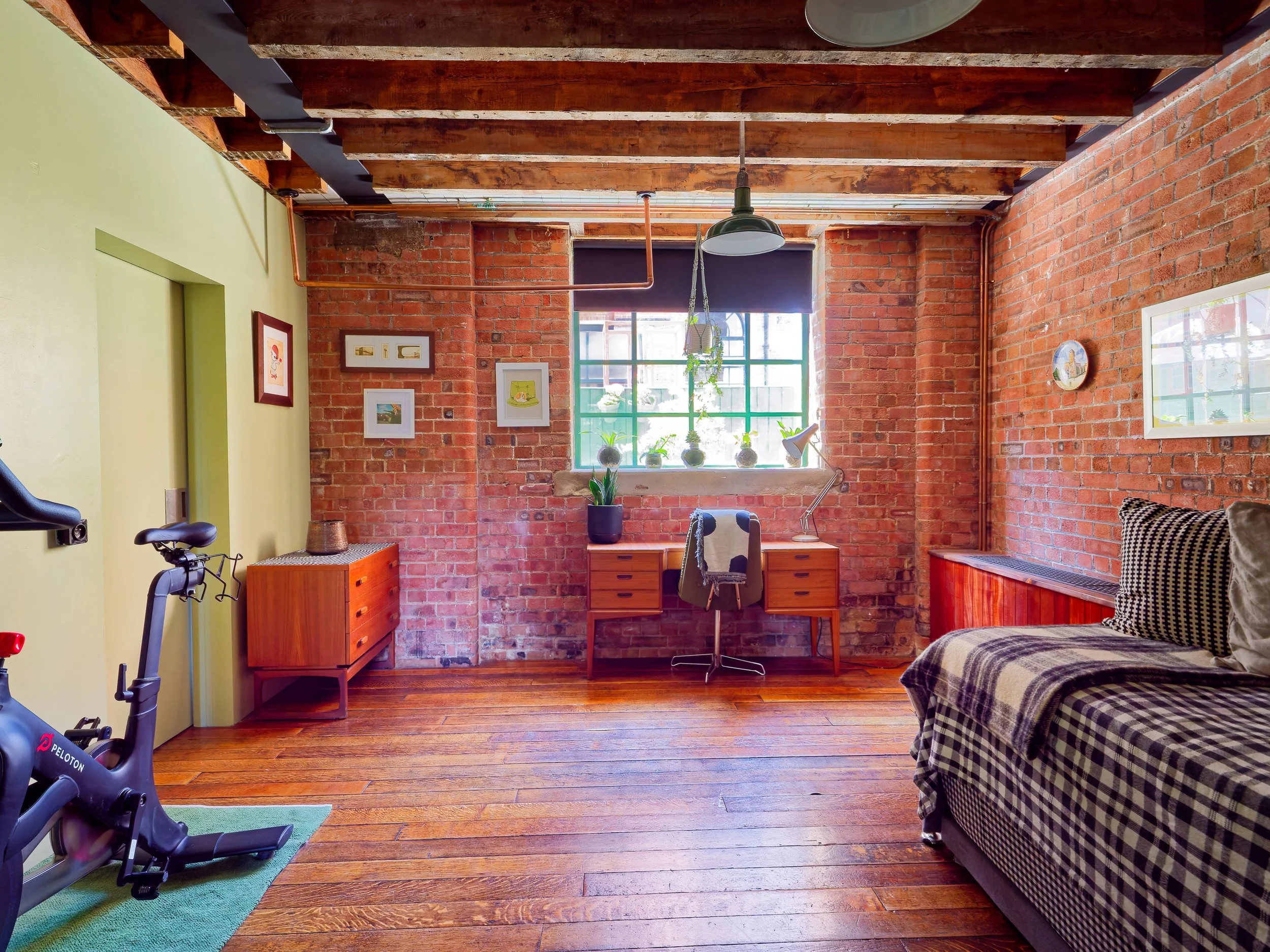 A cozy industrial-style living room with exposed brick walls, wooden ceiling beams, and hardwood floors. It features a desk with a chair near a window, a plaid couch, a small dresser, and a Peloton exercise bike. Decor includes framed artwork, small 