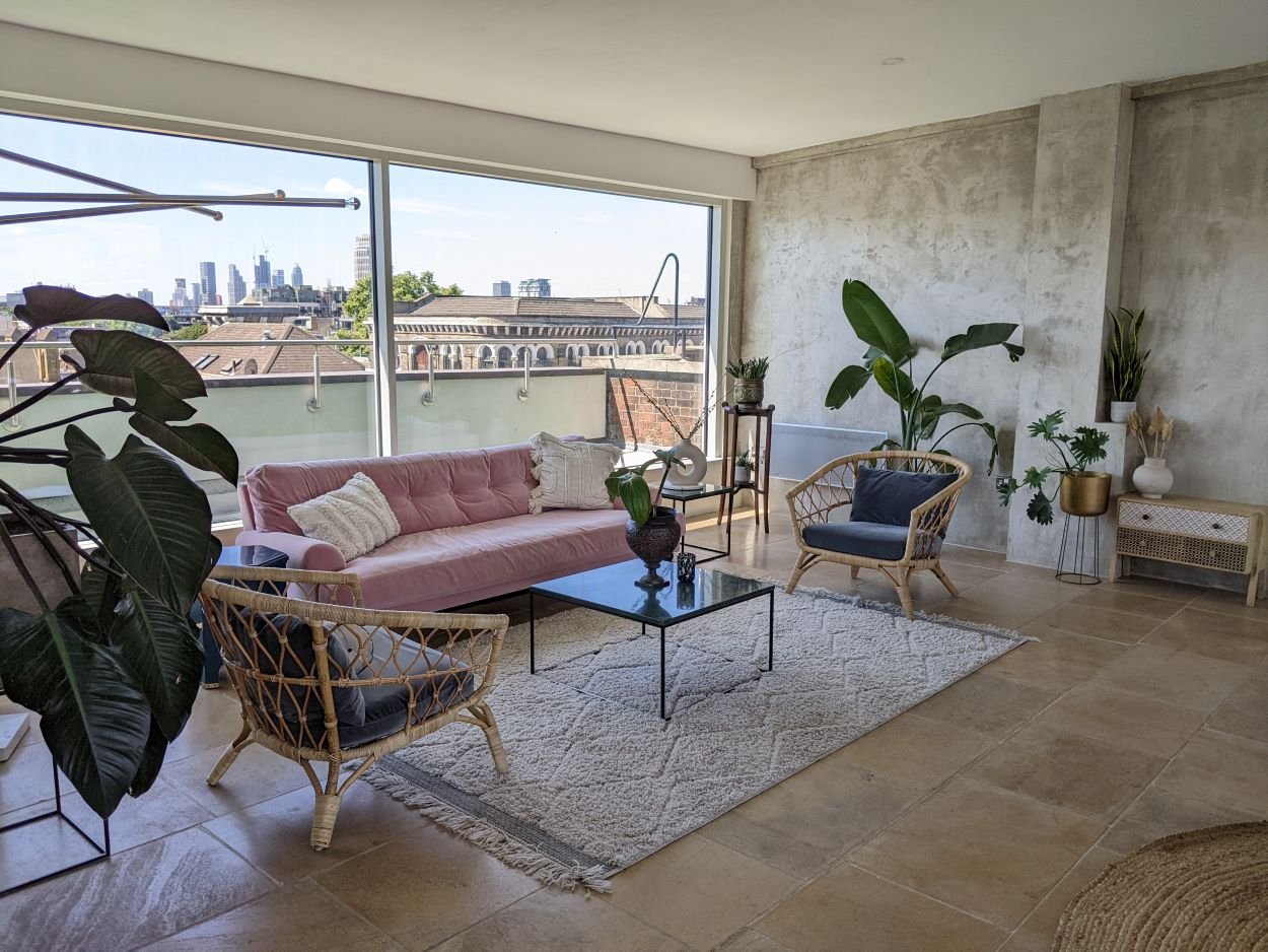 Living room with large window showing city skyline, pink sofa, rattan armchairs, potted plants, glass coffee table, white rug, and modern decor.