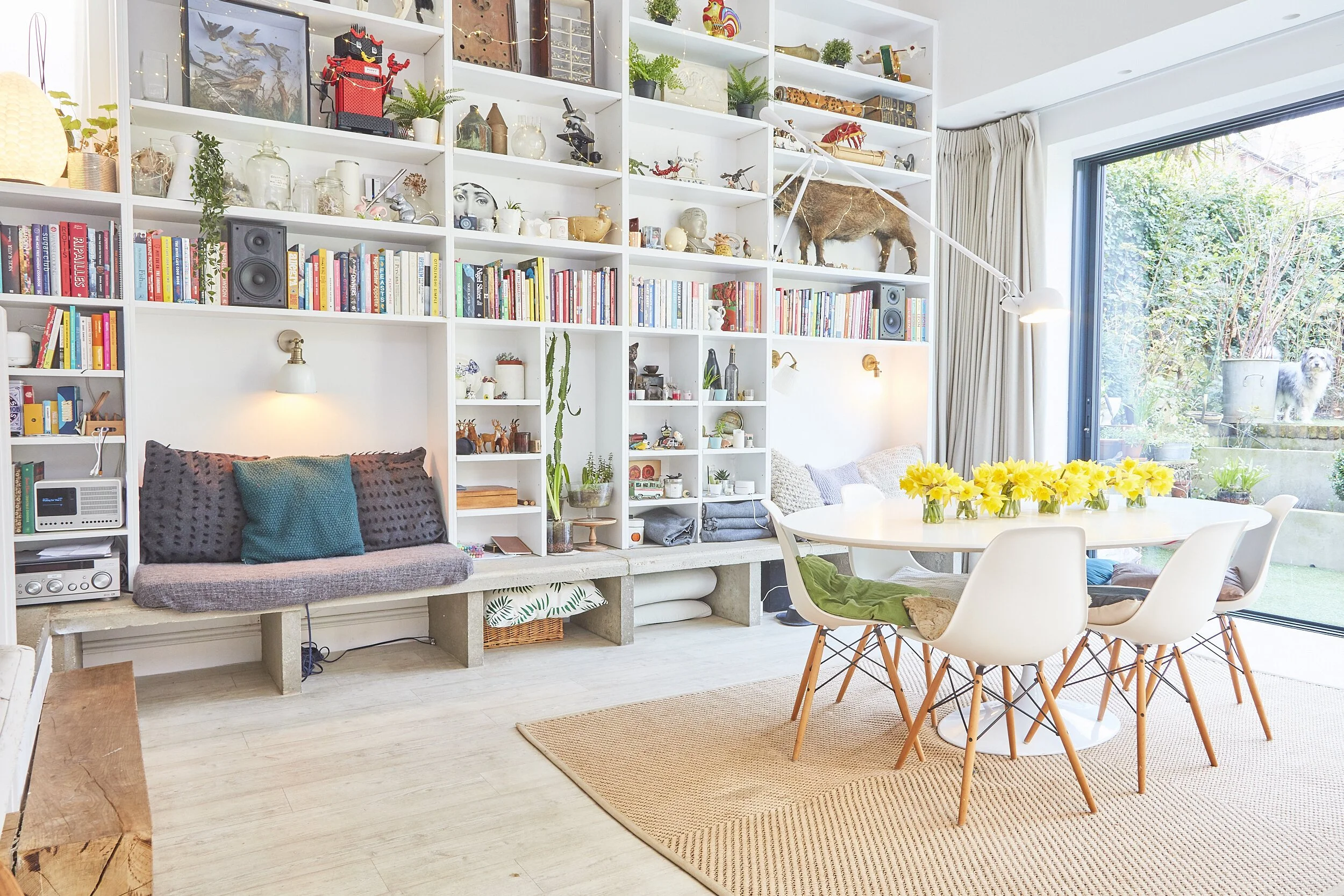 Bright dining and living room with a large window and a white bookshelf filled with books, plants, and decorative items, including animal figurines and art. There is a round dining table with white chairs and yellow flowers in a vase. The room has li