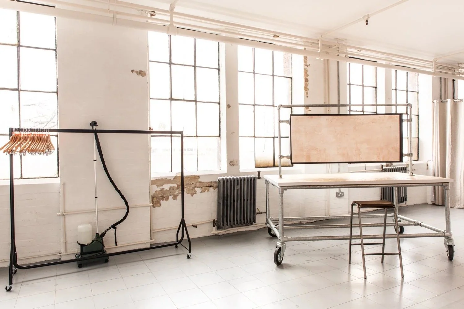 An industrial-style studio with large windows, white brick walls, and minimal furniture including a clothing rack with wooden hangers on the left, a metal table with a wooden panel and a wooden chair on wheels on the right, and vintage radiators belo