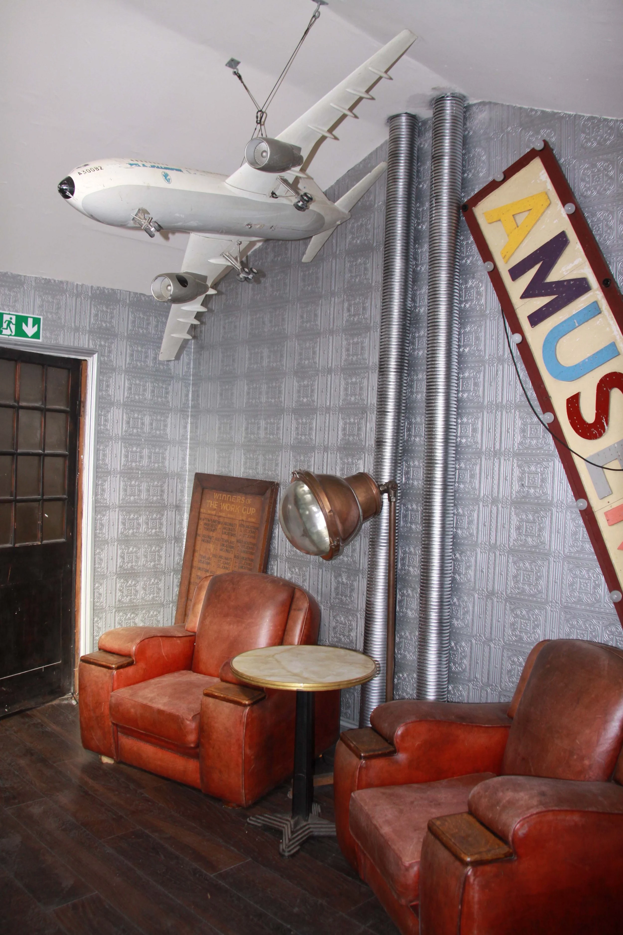 Interior with vintage aviation decor including a model airplane hanging from the ceiling, a large sign reading 'AMUSU', two brown leather armchairs, a small round table, a rust-colored badge or plaque, and metallic pipes on the wall.