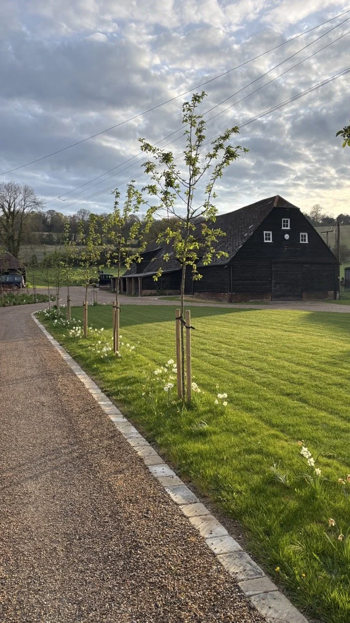 A paved pathway runs alongside a well-manicured grassy lawn with young trees planted along the edge, supported by wooden stakes, leading towards a large black barn with a steep roof under a partly cloudy sky.