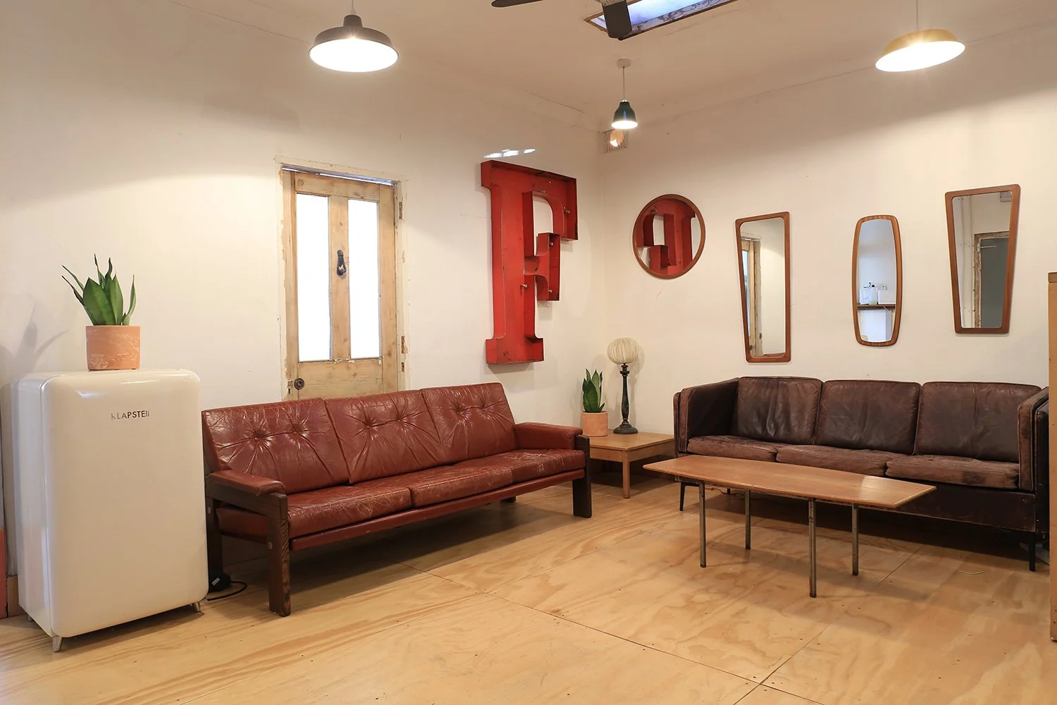 Living room with two vintage leather sofas, a wooden coffee table, a side table with a plant and a lamp, wall mirrors, a large red-letter sculpture, a potted plant, and an old door, with ceiling lights.