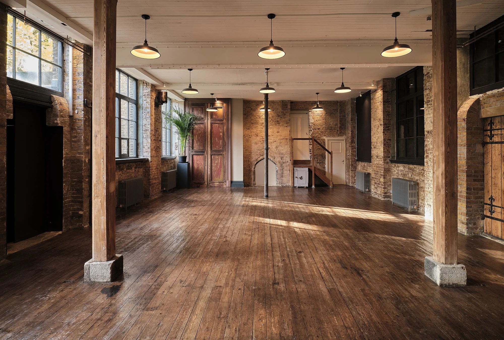 An empty loft-style room with exposed brick and wood beams, large windows, hardwood floors, and hanging pendant lights.
