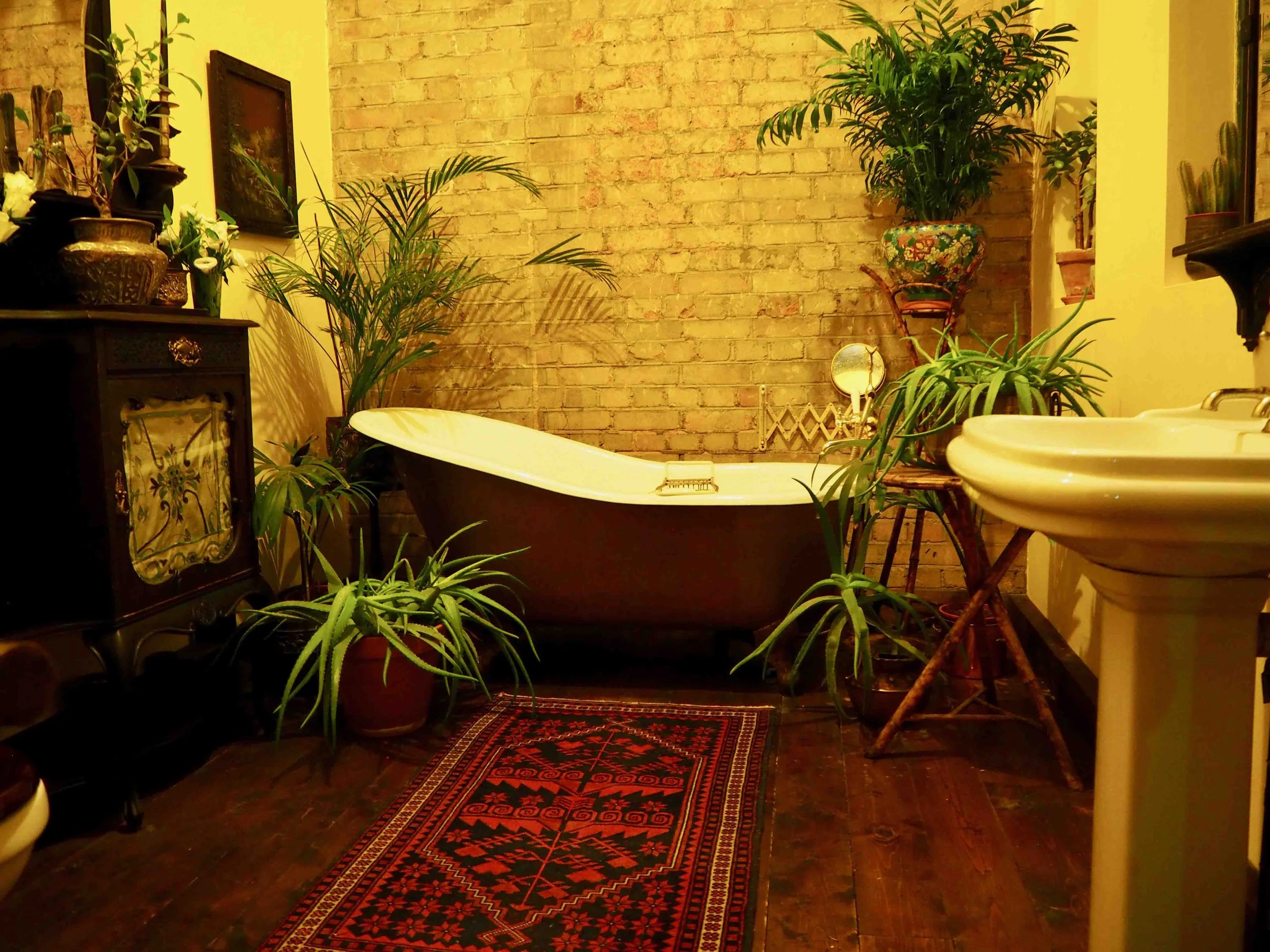 A vintage-style bathroom with a clawfoot bathtub surrounded by plants, a rug, an antique cabinet, a pedestal sink, and exposed brick wall.