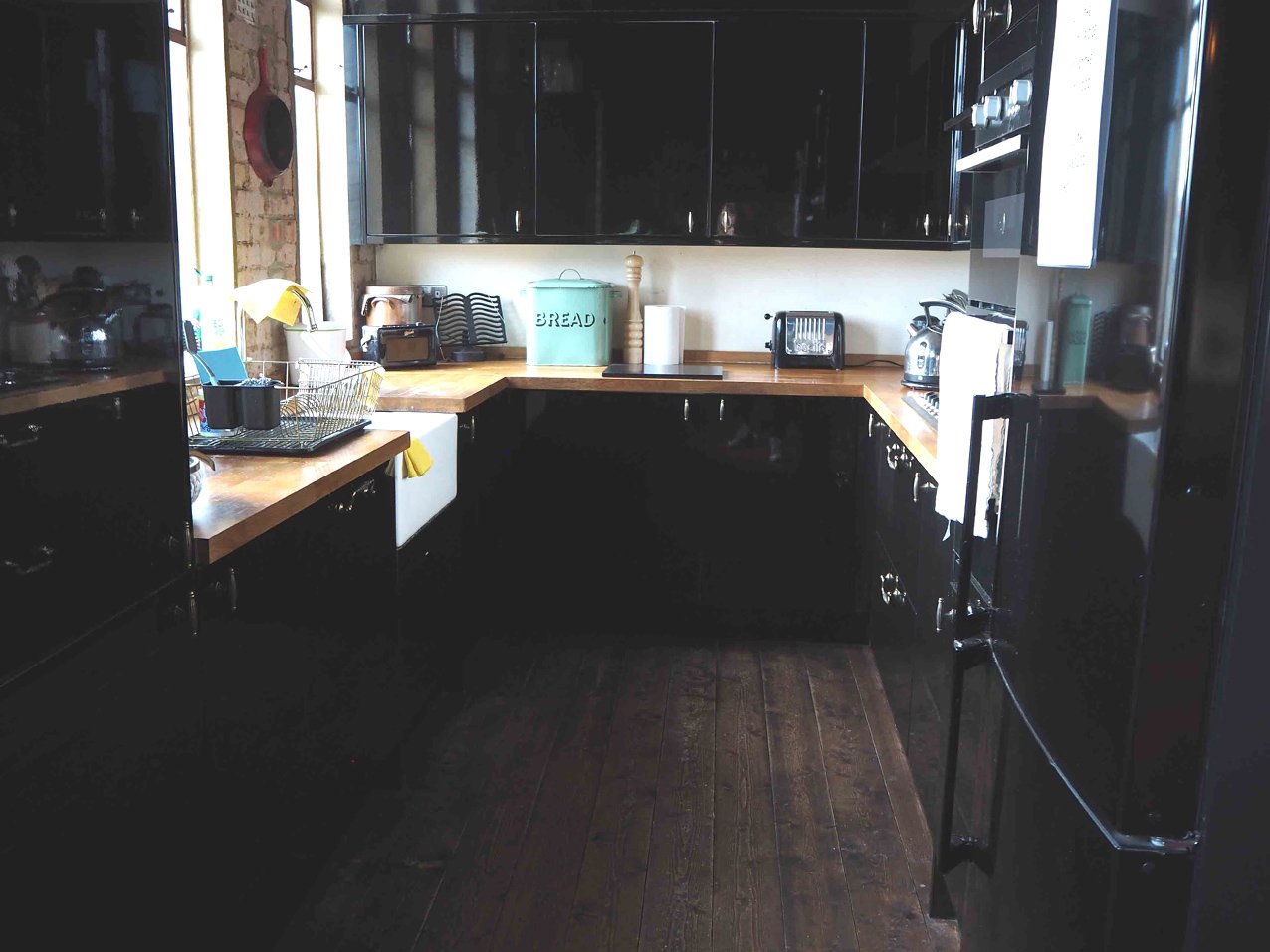 A kitchen with black cabinets and wooden countertops. There is a farmhouse sink, a dish drying rack, and various small appliances including a toaster and kettle. A bread bin, paper towel roll, and some dishes are on the counter. There's a window with