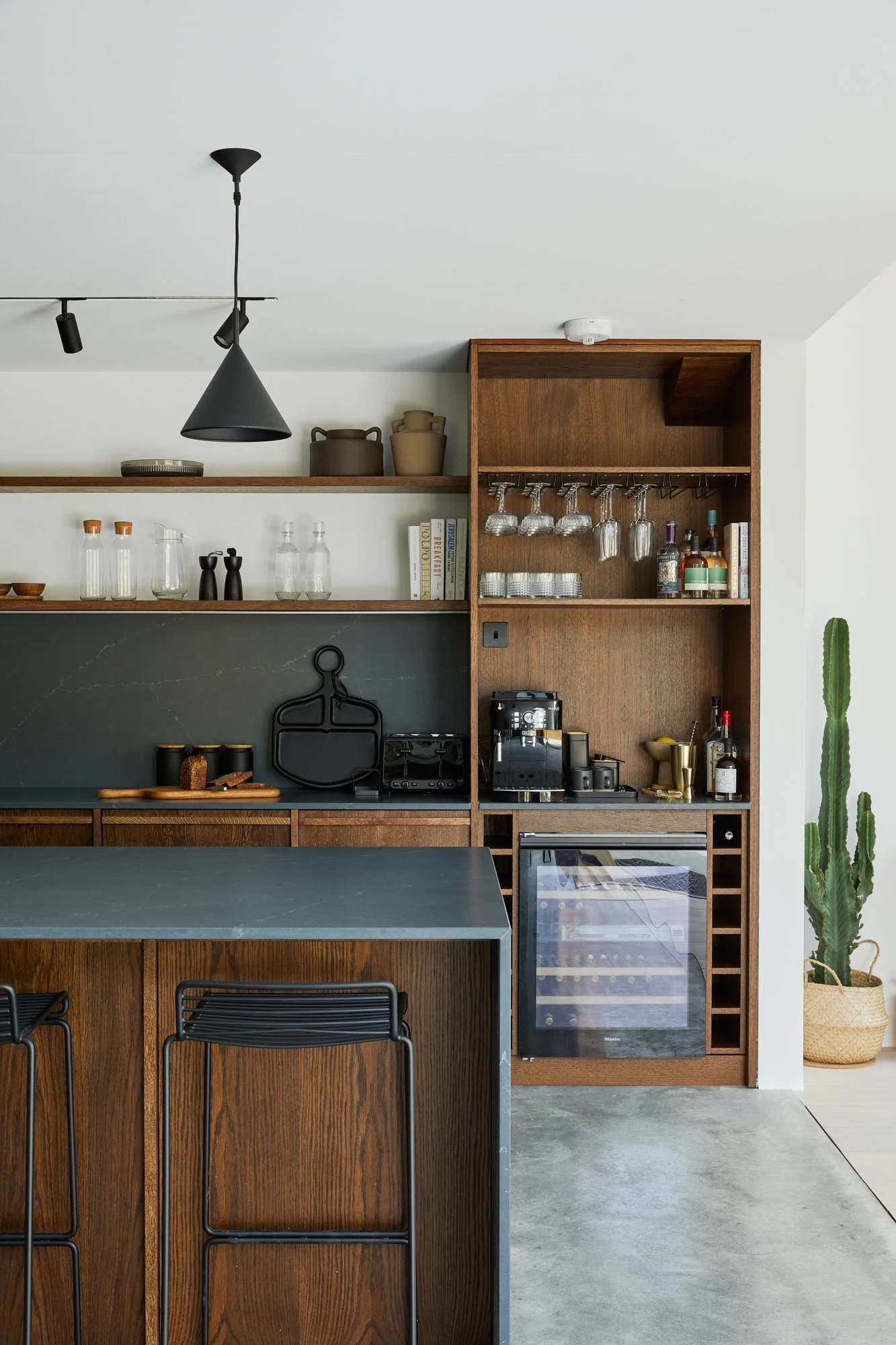 Modern kitchen with wooden cabinetry, open shelving, a small wine fridge, bar stools, and a potted cactus.