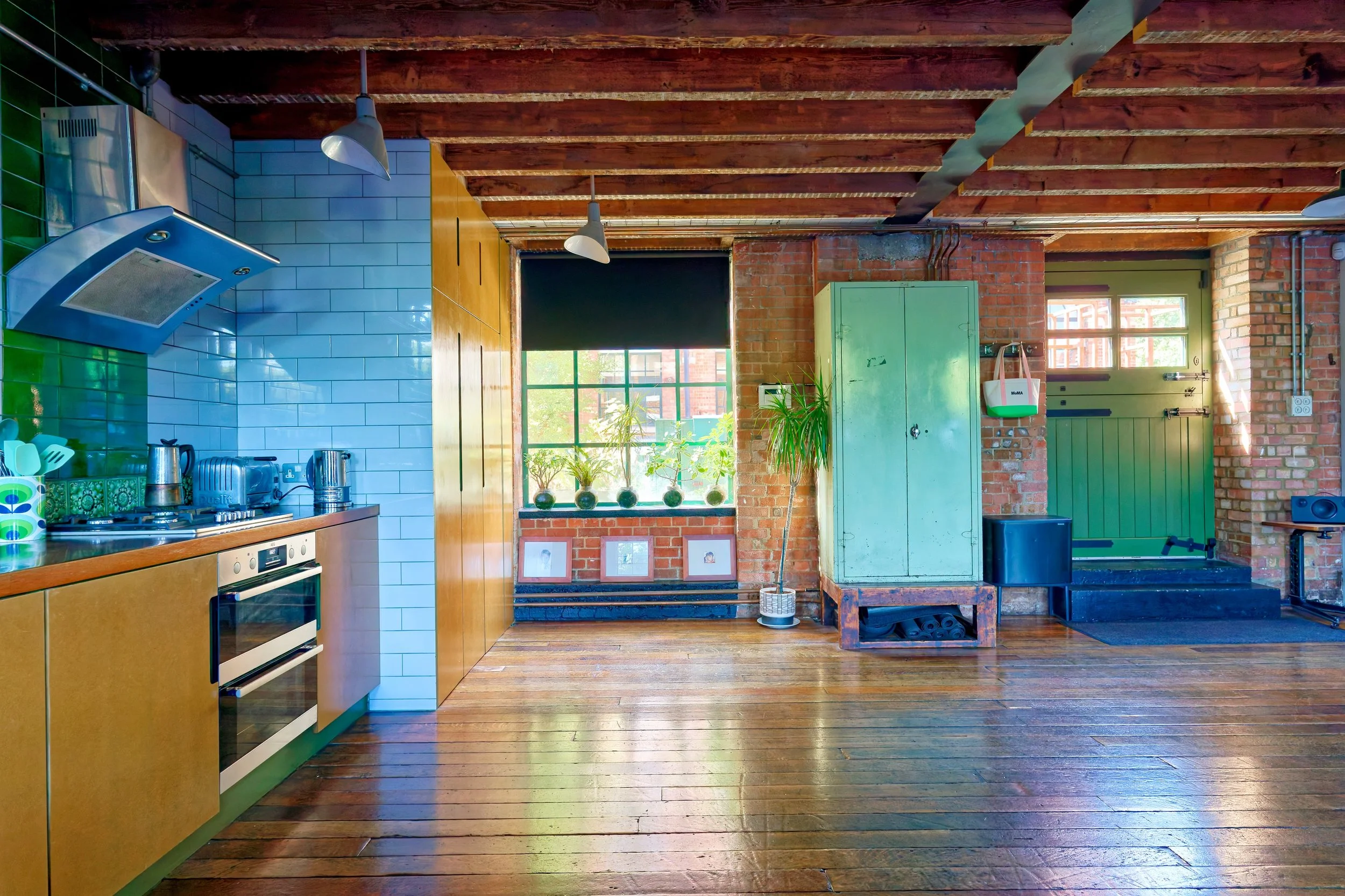 Open kitchen space with wooden floors, brick walls, a window with plants, a green door, and industrial-style ceiling beams.