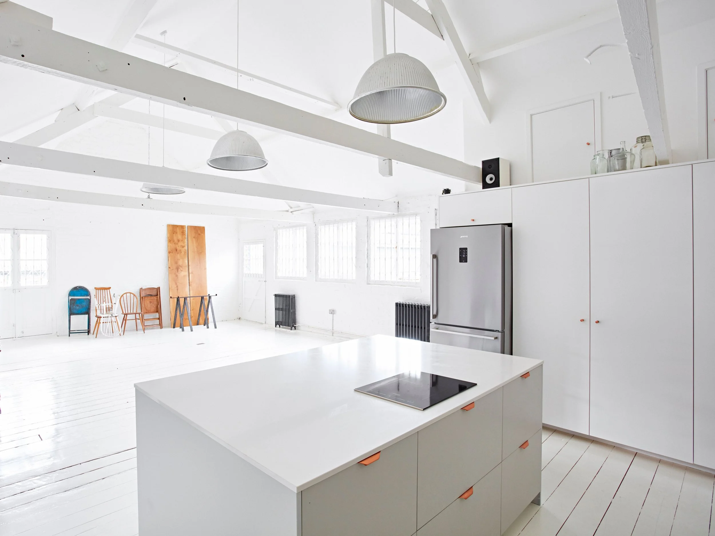 Bright white kitchen with a island, refrigerator, and white cabinets. Several chairs are arranged against the wall in an adjacent room.