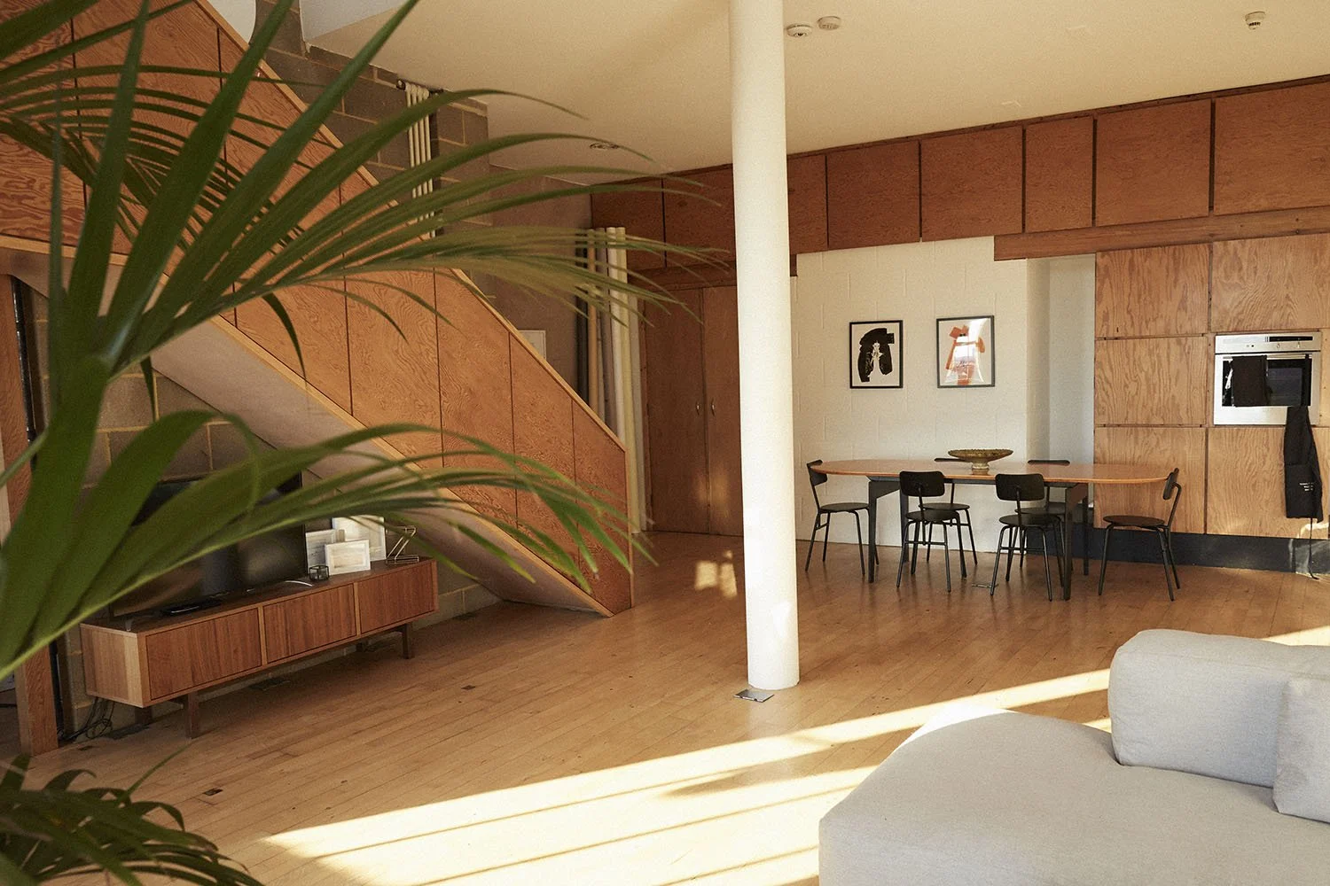 Modern living room with wooden flooring, a white sofa, a wooden TV console, and a staircase with wooden paneling. There is a round dining table with six black chairs, framed artwork on the wall, and a white column in the space. Sunlight streams in, c