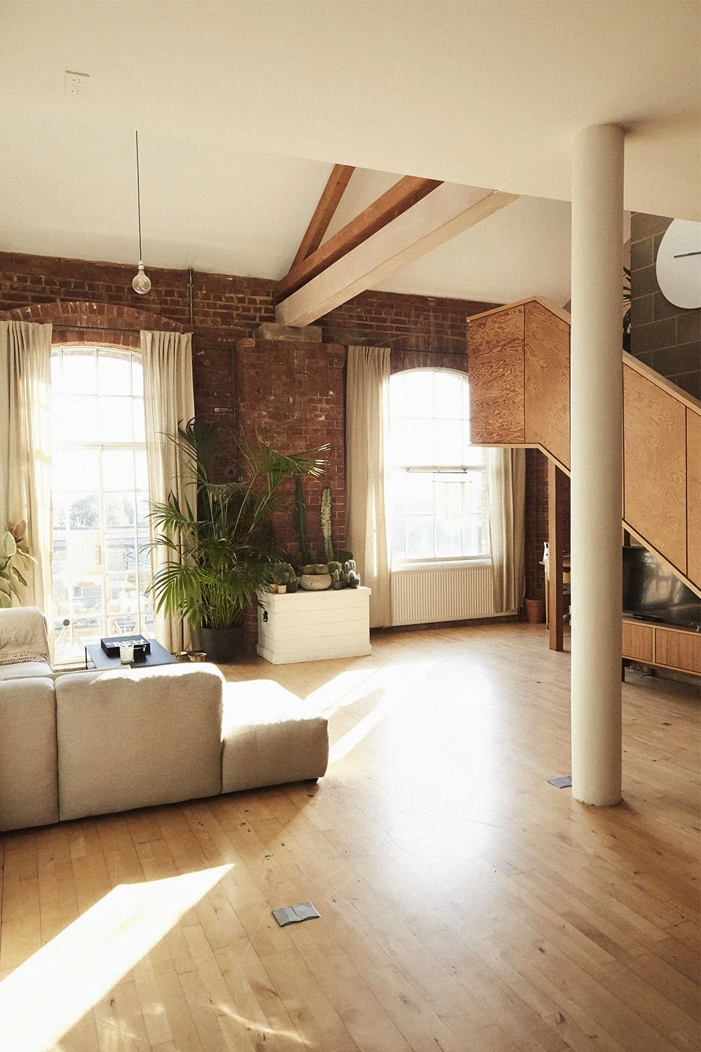Bright living room with large windows, exposed brick walls, a white sofa, potted plants, wooden staircase, and hardwood floors.