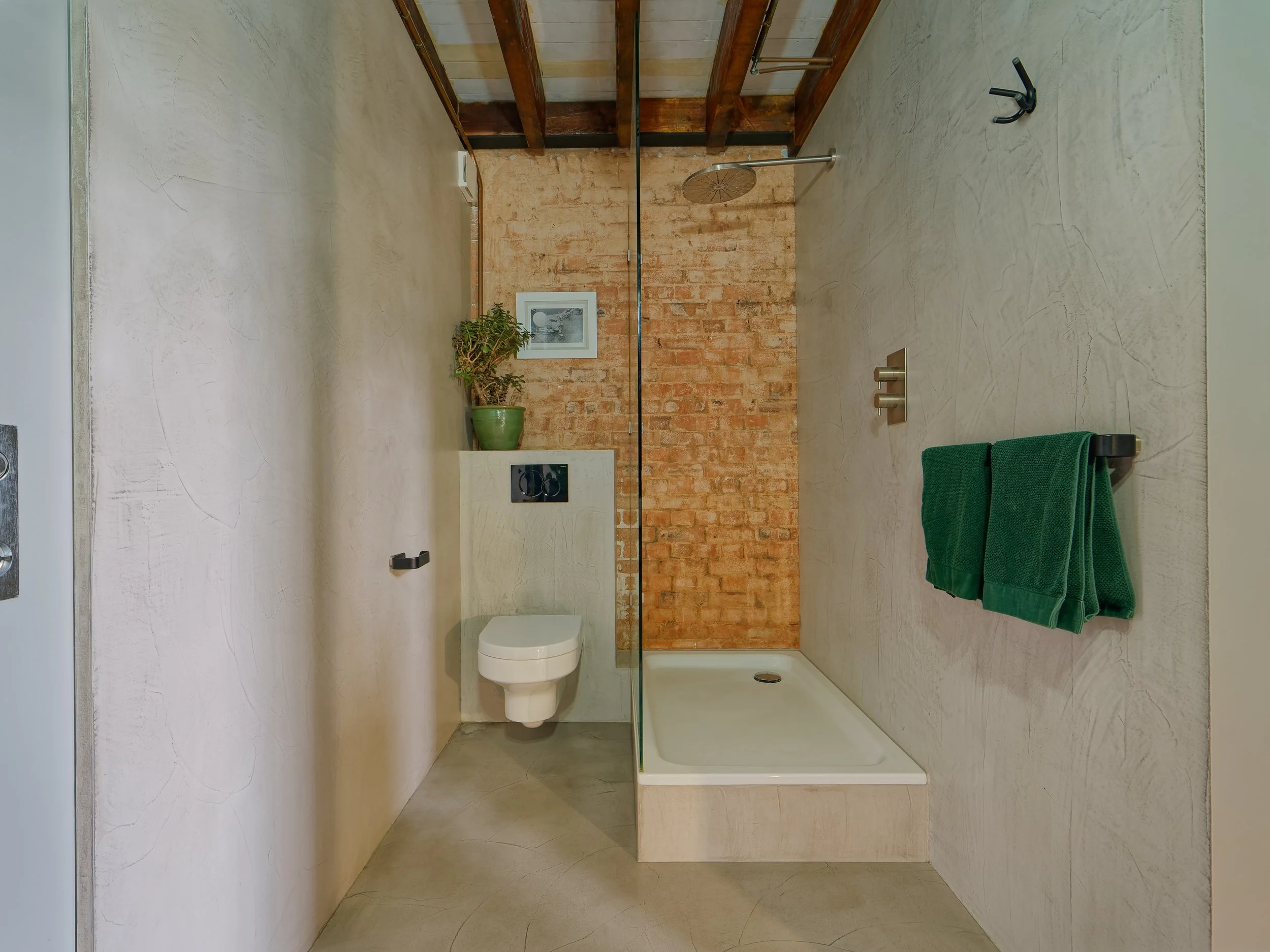 Bathroom with beige textured walls, a brick accent wall in the shower area, a shower head, green towels on a rail, a potted plant, framed picture, and a white toilet.