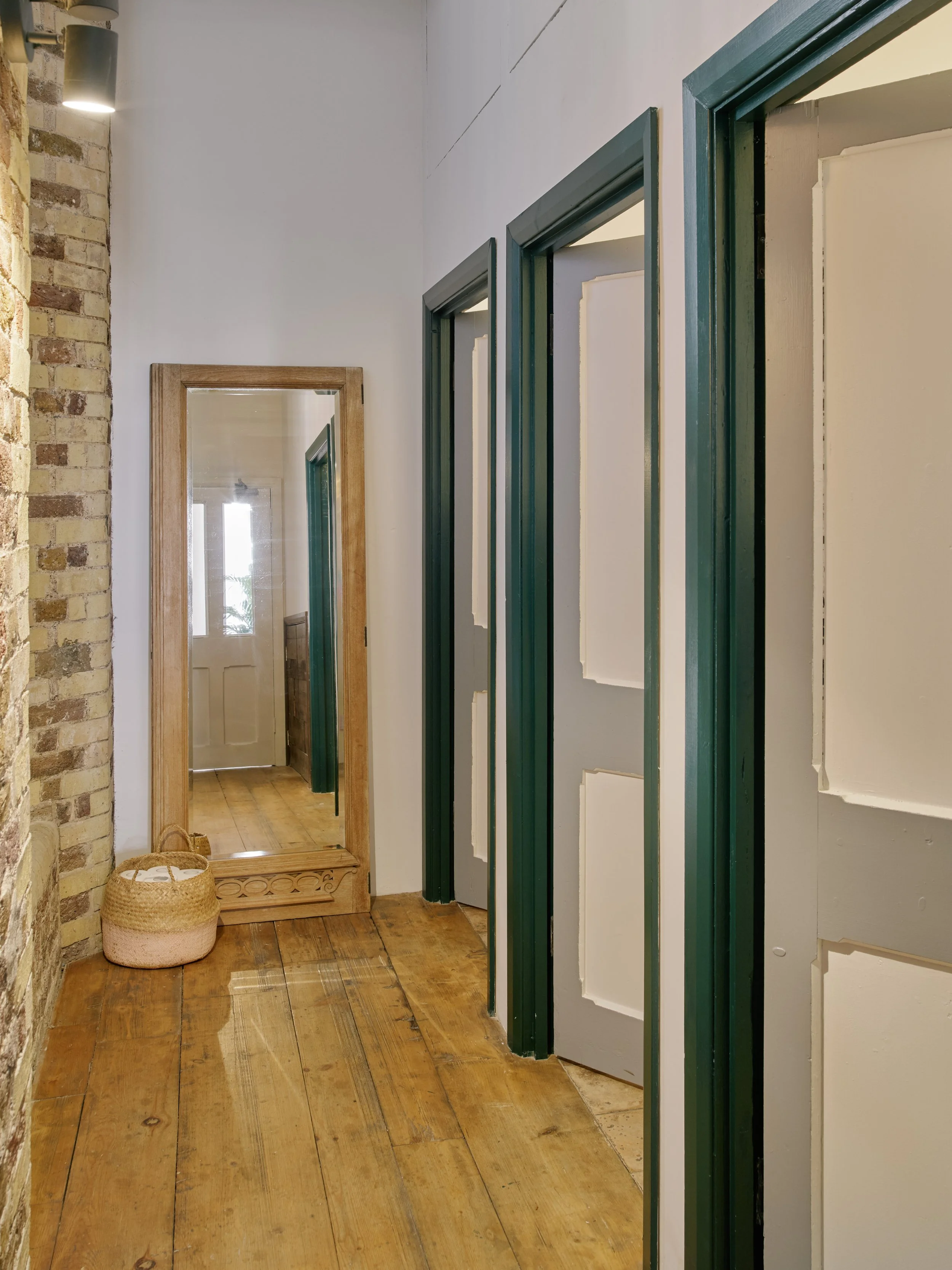 Hallway with wooden floor, brick wall on the left, white walls, a tall mirror on the left, and open doors leading to rooms on the right.
