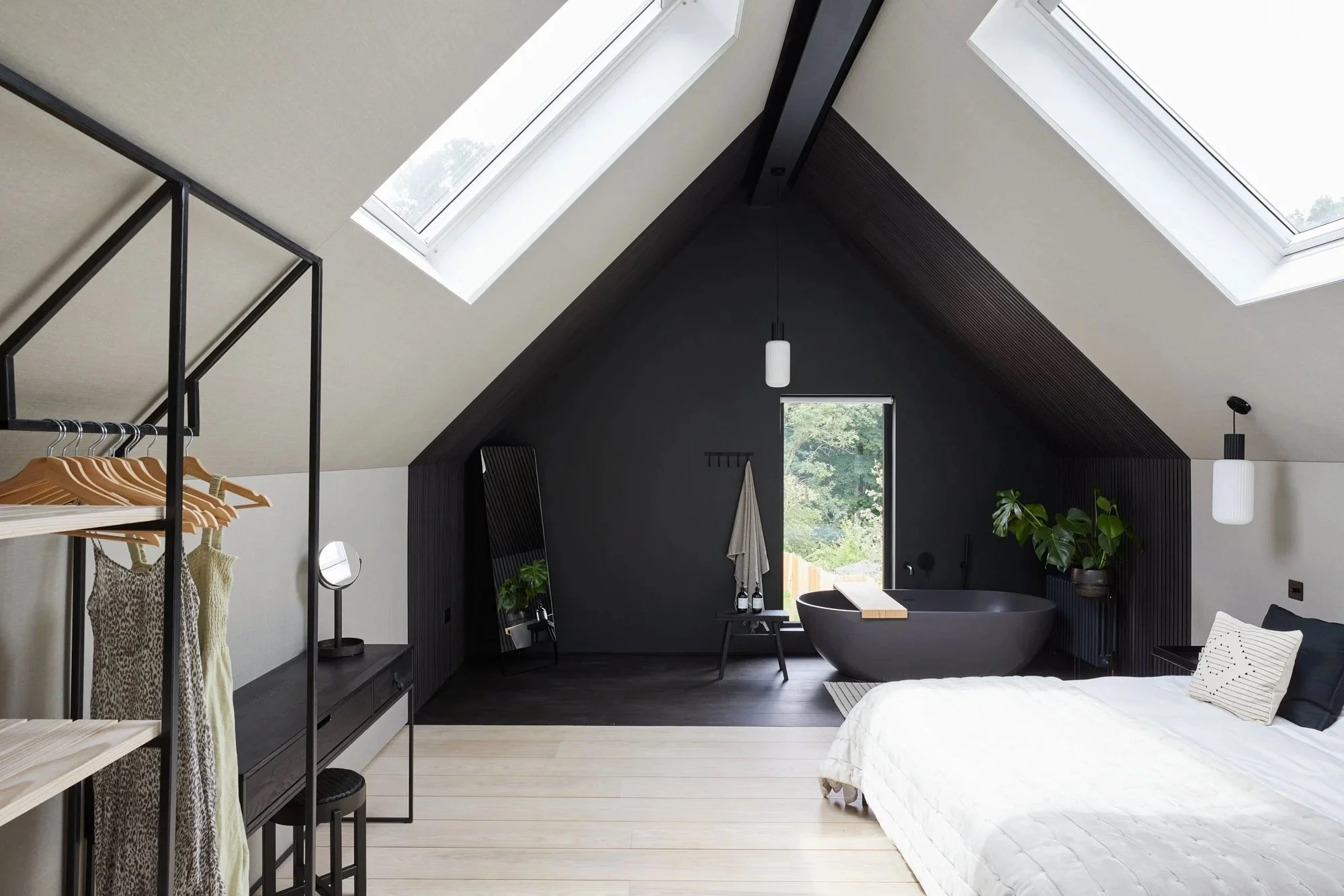 A modern bedroom with a sloped ceiling featuring skylight windows, a black bathtub near a window, a large bed with white bedding, and minimalistic decor including plants and a clothing rack.