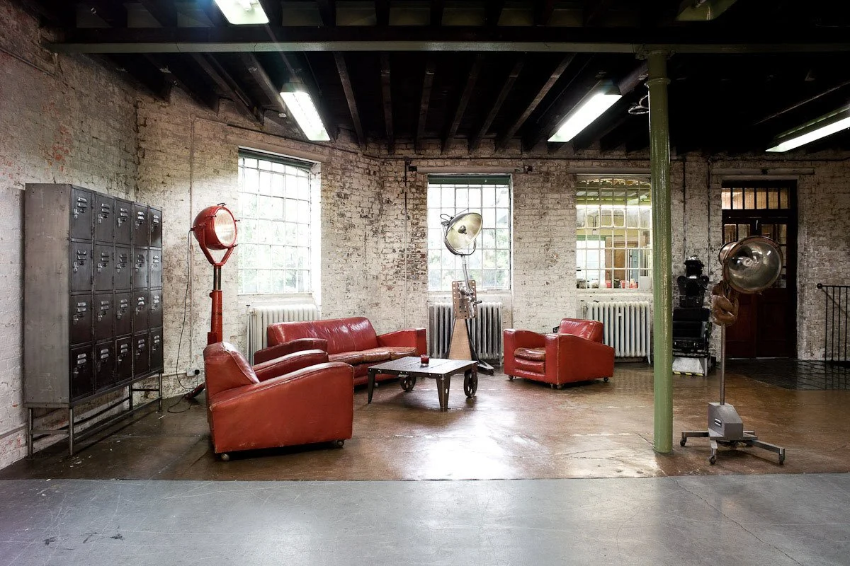 Industrial lounge with brick walls, large windows, red leather sofas, vintage lighting, lockers, and photography equipment.