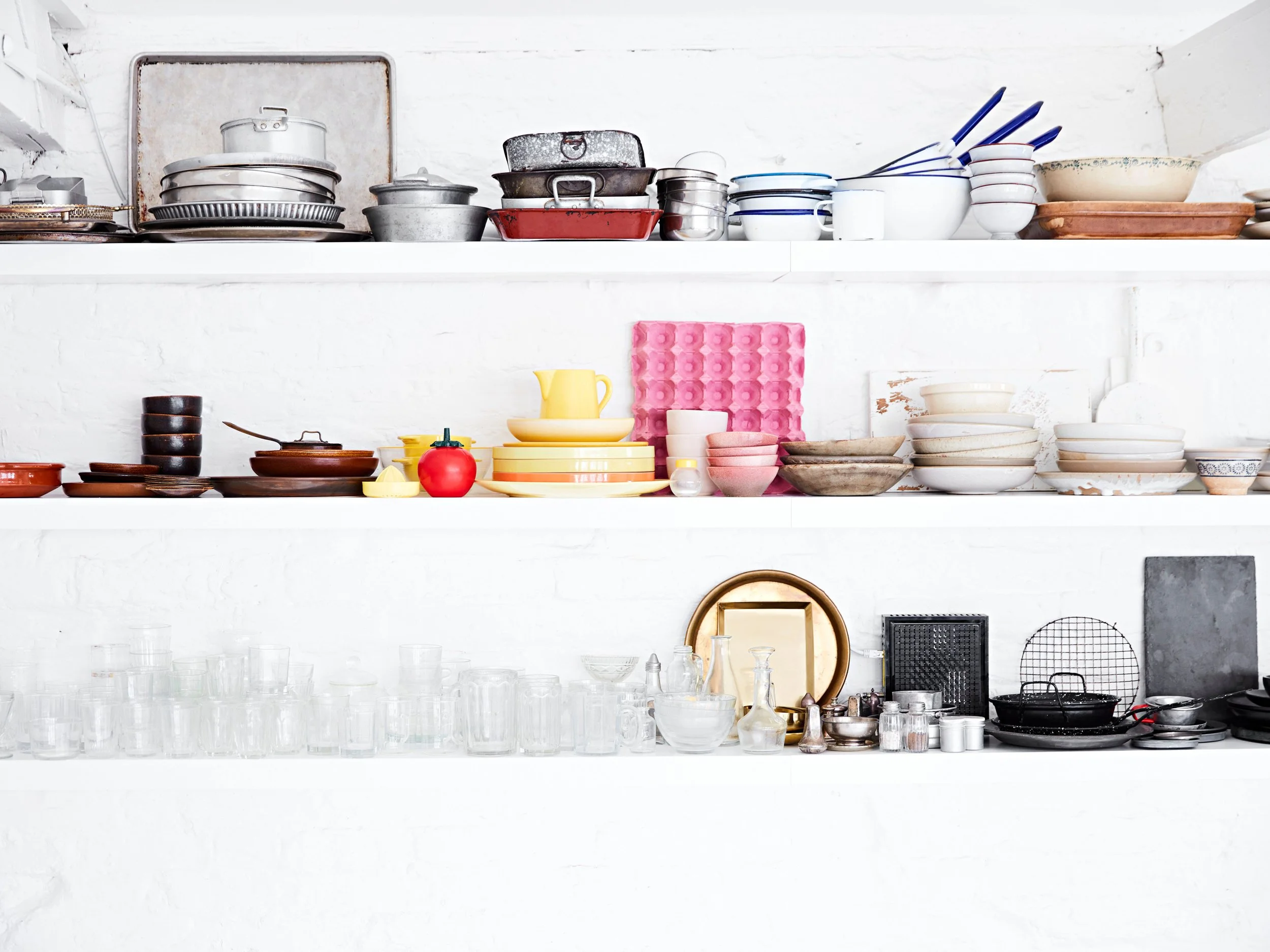 Open kitchen shelves filled with dishes, bowls, plates, pots, and kitchenware in various styles and colors against a white brick wall.