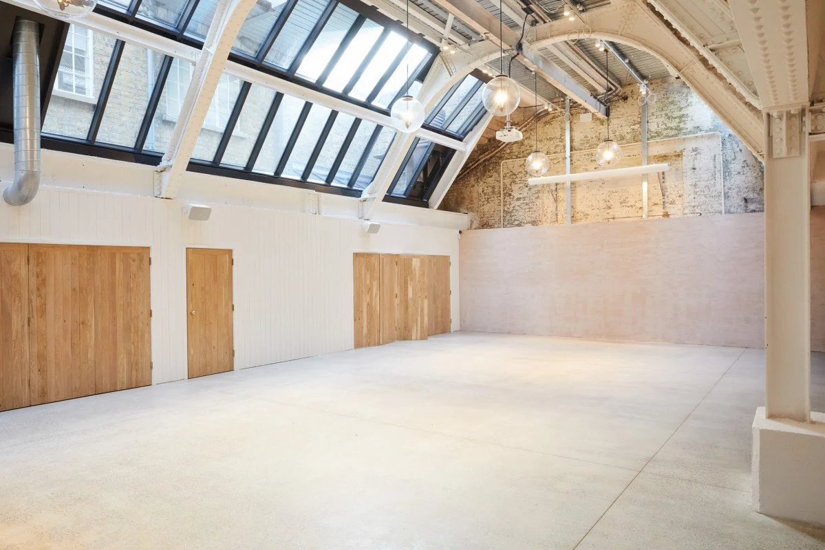 A spacious empty room with large skylight windows, exposed beams, hanging globe lights, a mix of exposed brick and smooth painted walls, and a polished concrete floor.