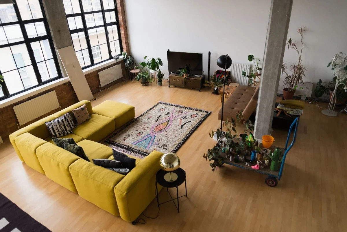 A spacious living room with large windows, a yellow sectional sofa, a colorful area rug, a flat-screen TV on a stand, and houseplants scattered around. There is also a brown couch, a rolling cart with plants, and a small side table with a lamp.