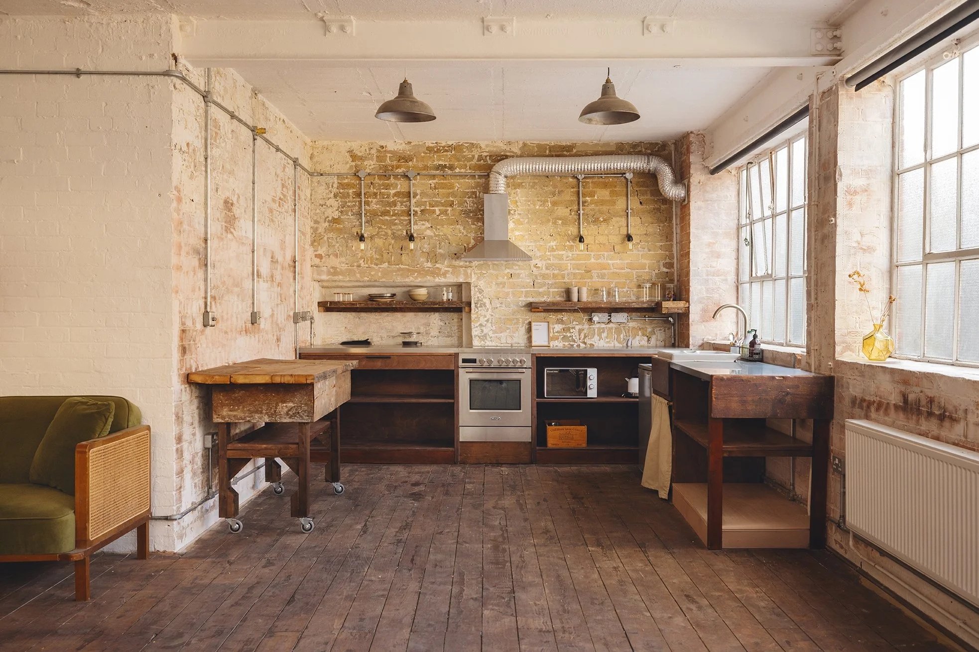 An industrial-style kitchen with exposed brick walls, large windows, wooden floors, and minimalist open shelving. The kitchen features a stove, microwave, and sink, with pendant lights hanging from the ceiling.
