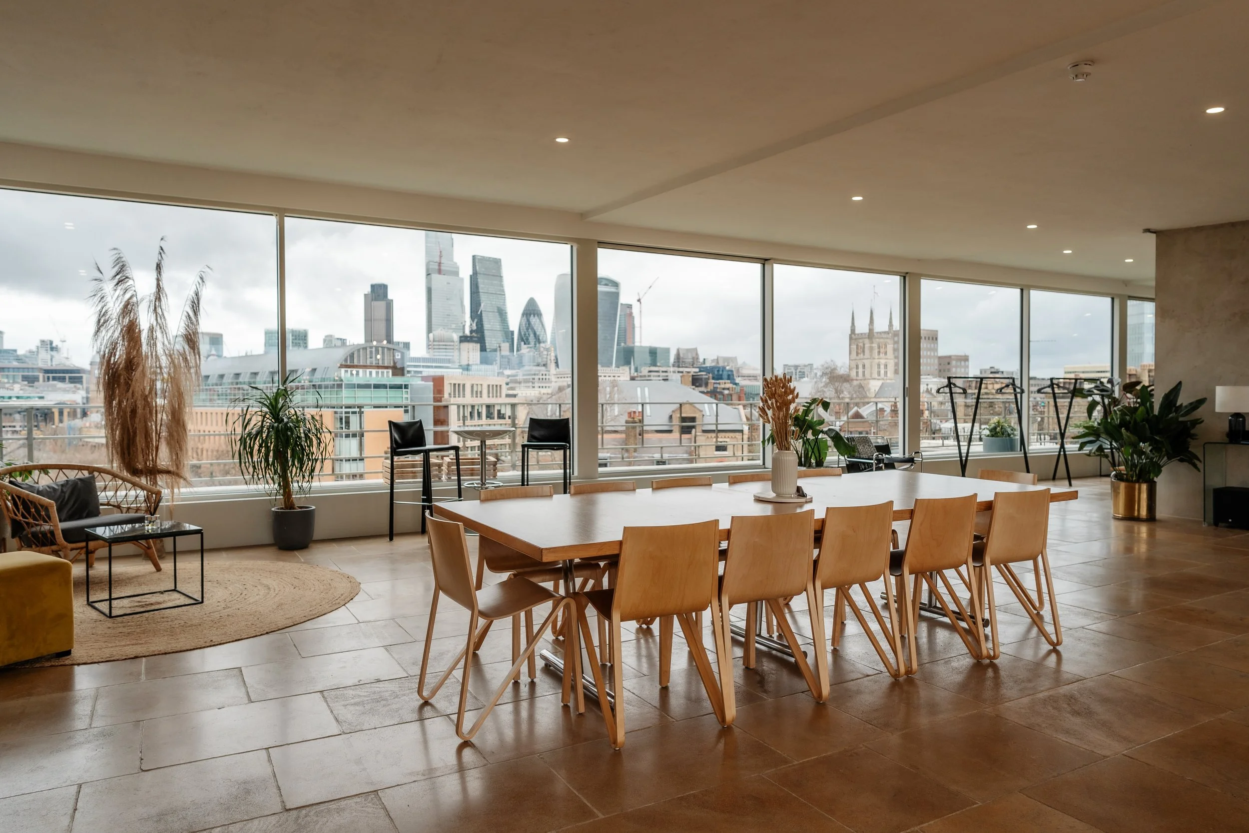 Modern conference room with large windows overlooking a city skyline, including skyscrapers and historic buildings, with a long wooden table, chairs, indoor plants, and a seating area.