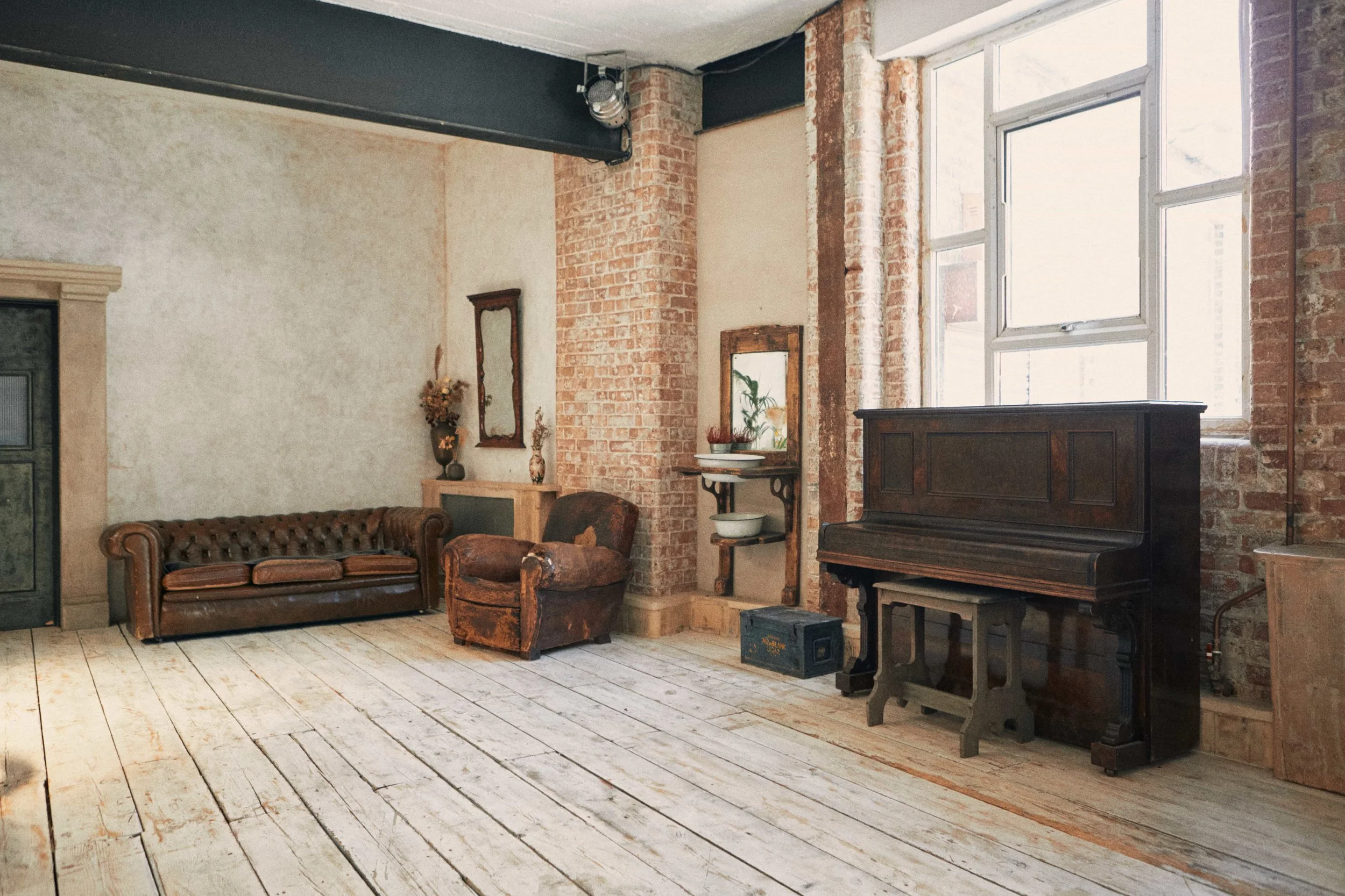 Vintage living room with exposed brick walls, distressed wooden floor, dark leather sofa and armchair, antique piano, and large window