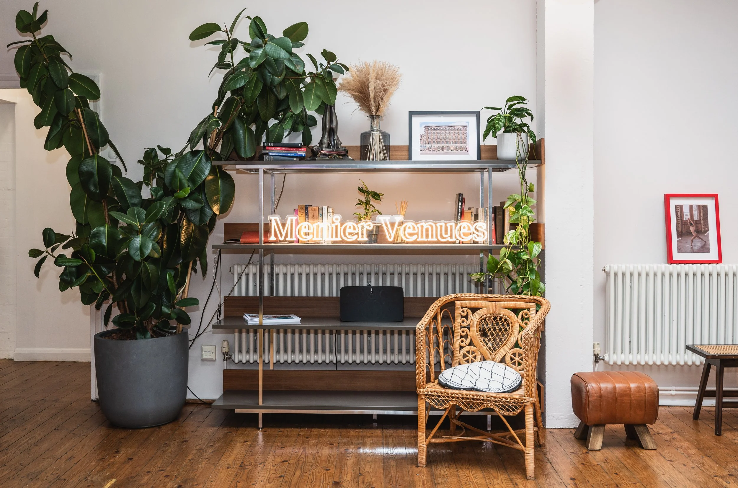 Interior of a room with a tall potted plant on the left, a shelf with decorative items and books, a neon sign that says 'Menton Venues,' a wicker chair with a cushion, a small leather ottoman, and framed artwork on the wall.