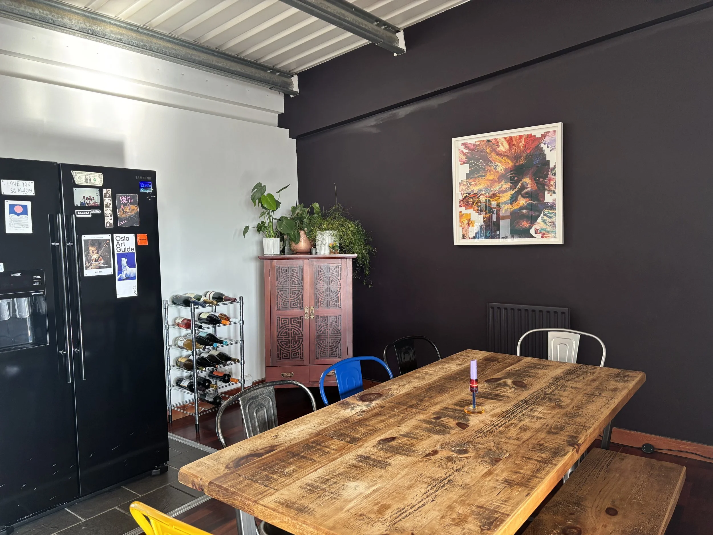 Interior of a room with a wooden dining table, metal chairs, a black refrigerator, a small wine rack, a pink cabinet with plants on top, a colorful abstract portrait on a dark wall, and a radiator.