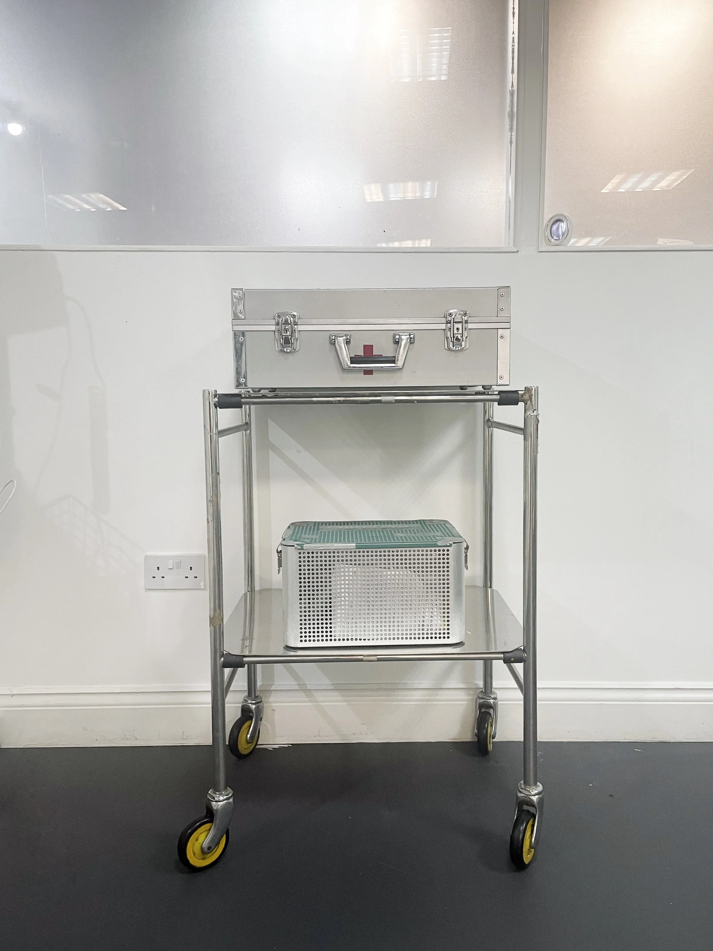 A medical trolley with a metal first aid kit on top and a perforated metal container on the lower shelf, against a white wall with a frosted glass window above.