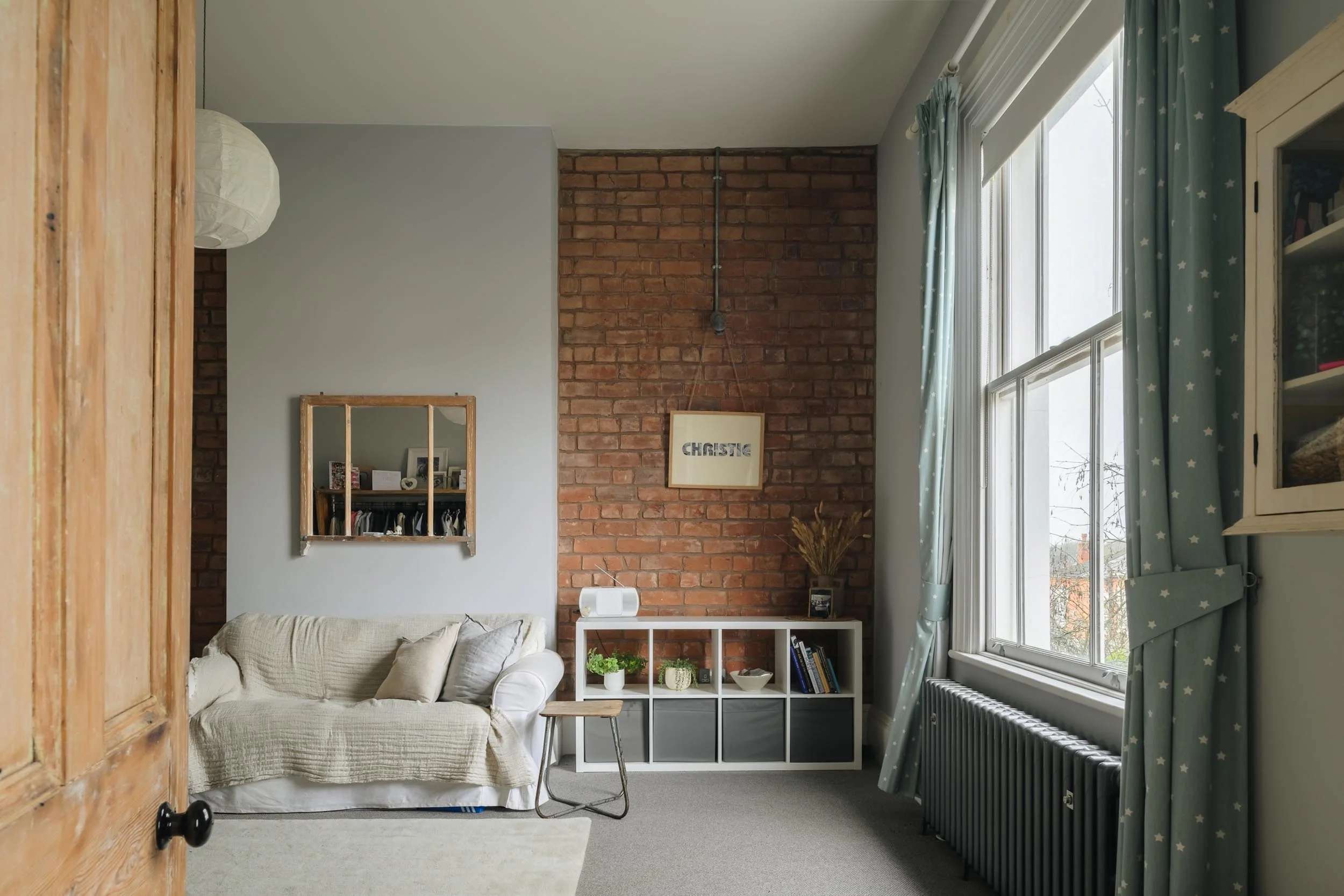 Living room with a white sofa, a small wooden stool, a white shelf with decor, a brick accent wall with framed art, large window with star-patterned curtains, and a radiator.