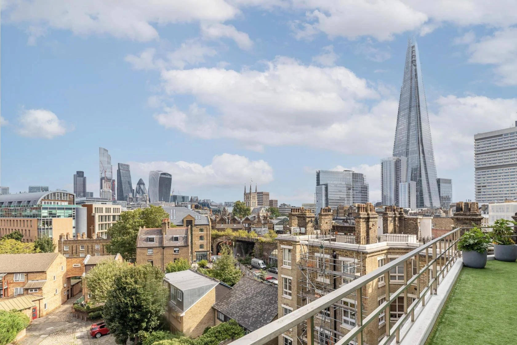Cityscape of London with modern skyscrapers like The Shard among older buildings, viewed from a rooftop terrace with potted plants and green artificial grass, under a partly cloudy sky.
