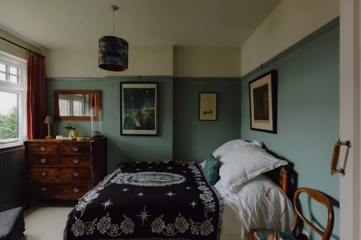 A bedroom with a window, a wooden dresser with a plant and lamp, a bed with white sheets and a black bedspread, framed artwork on the walls, and a wooden chair.
