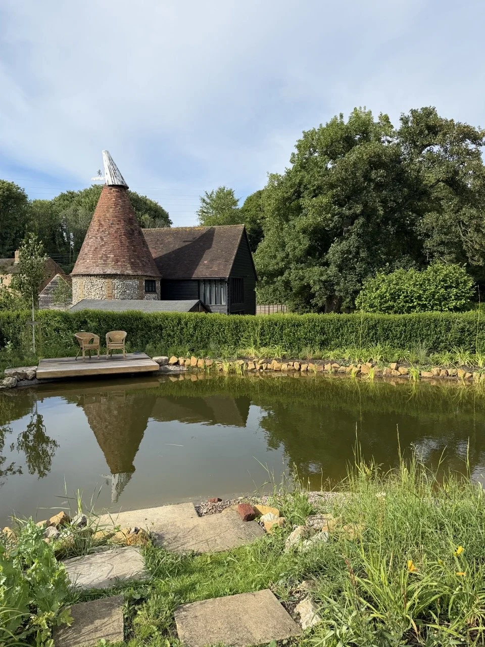 A pond with a small wooden dock and two chairs, surrounded by grass, rocks, trees, and a small house with a cone-shaped roof in the background.