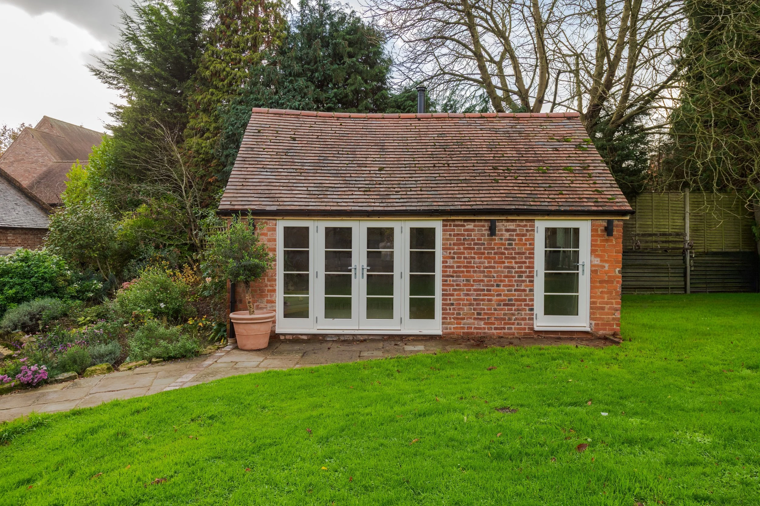 Small brick garden building with white-framed glass doors and windows, set in a green backyard with a paved patio and surrounded by trees and shrubs.